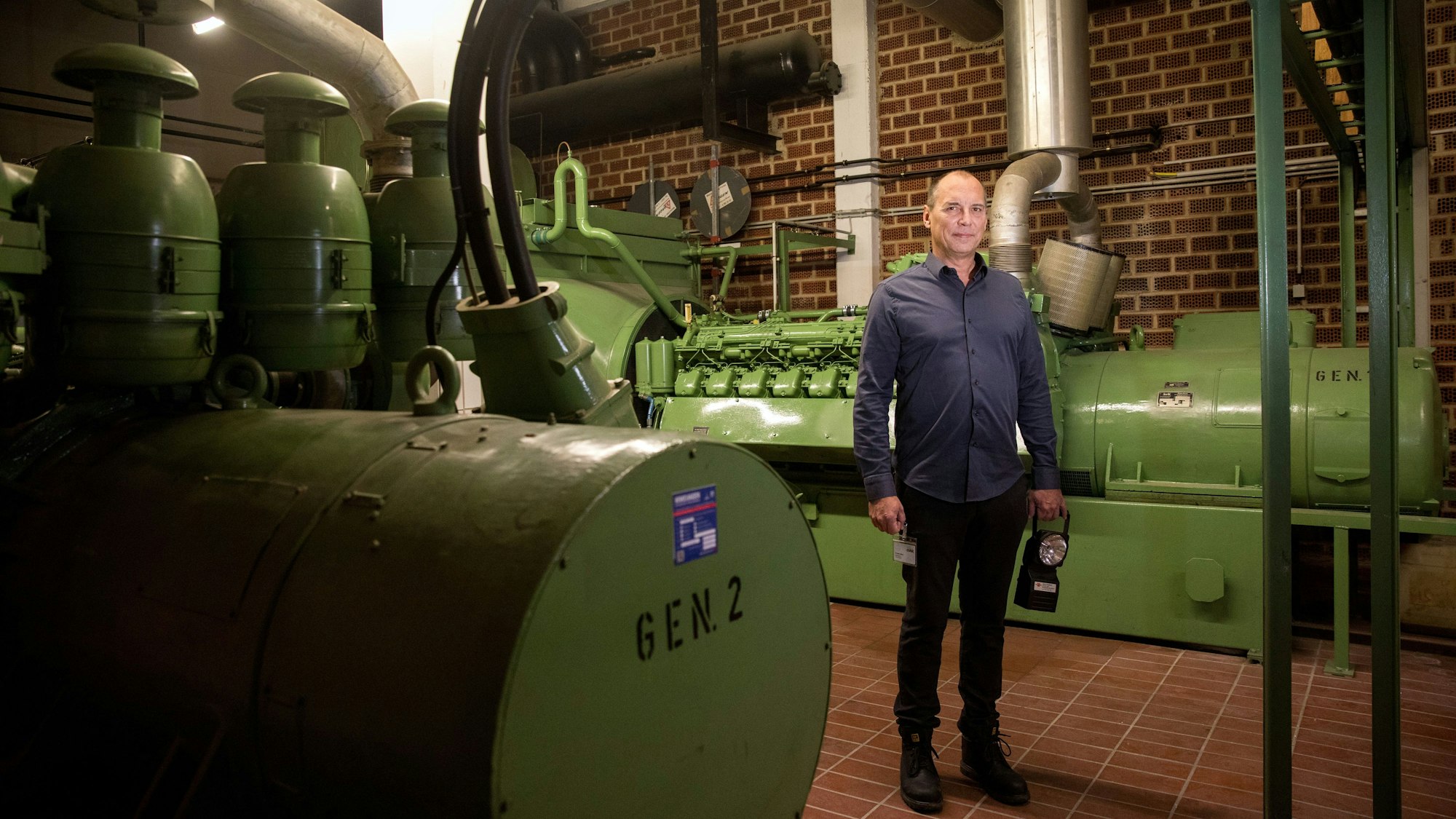 Blick in den Raum mit den Notstromaggregaten: Die großen grünen Tanks füllen den Raum mit roten Backsteinwänden und -boden, Thomas Velten steht dazwischen.