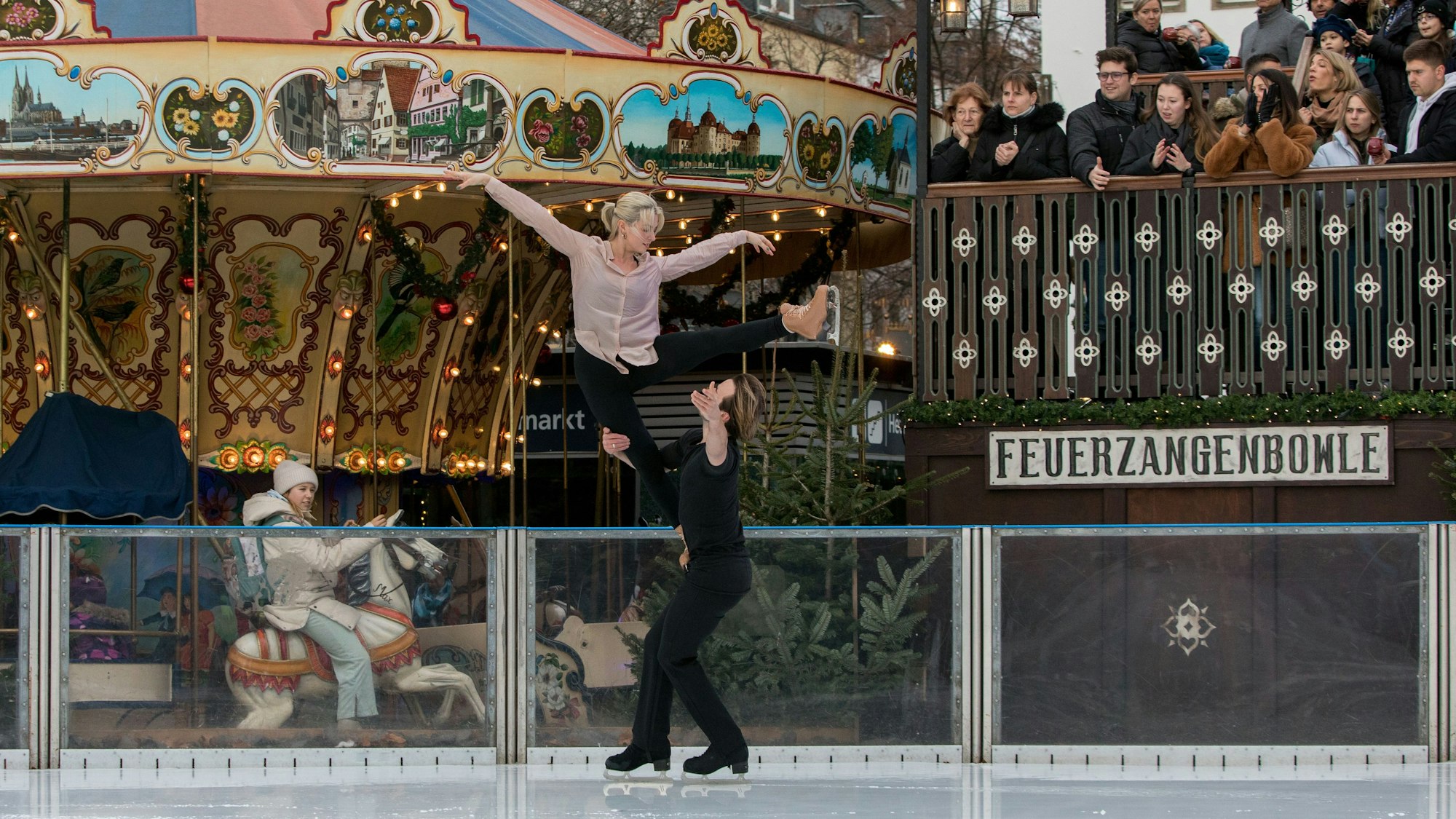 Tim Dieck hebt Olivia Smart über seinen Kopf, sie streckt die Arme aus, im Hintergrund ein Kinderkarussell des Weihnachtsmarkts auf dem Heumarkt.