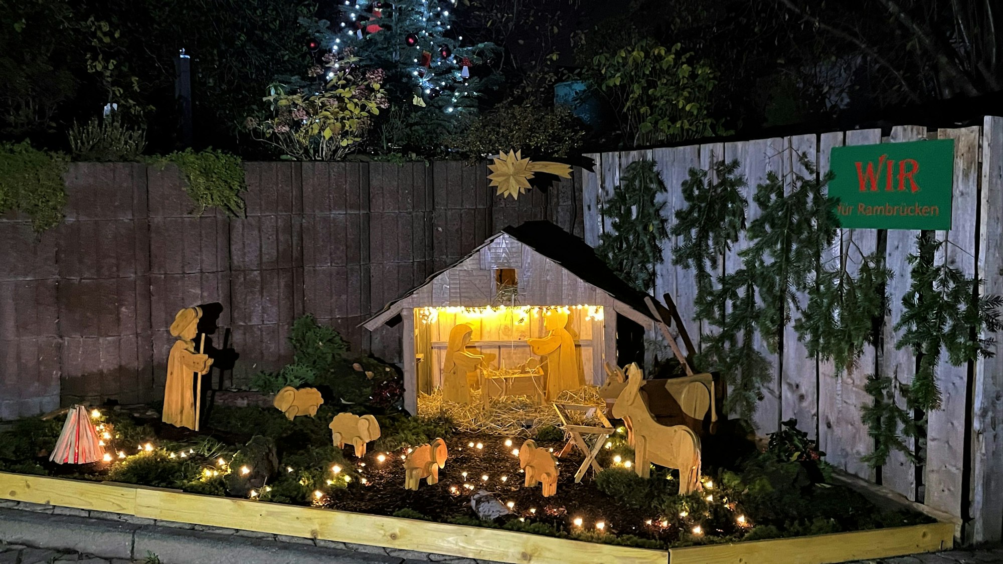 Eine Holzkrippe steht vor einem beleuchteten Tannenbaum.