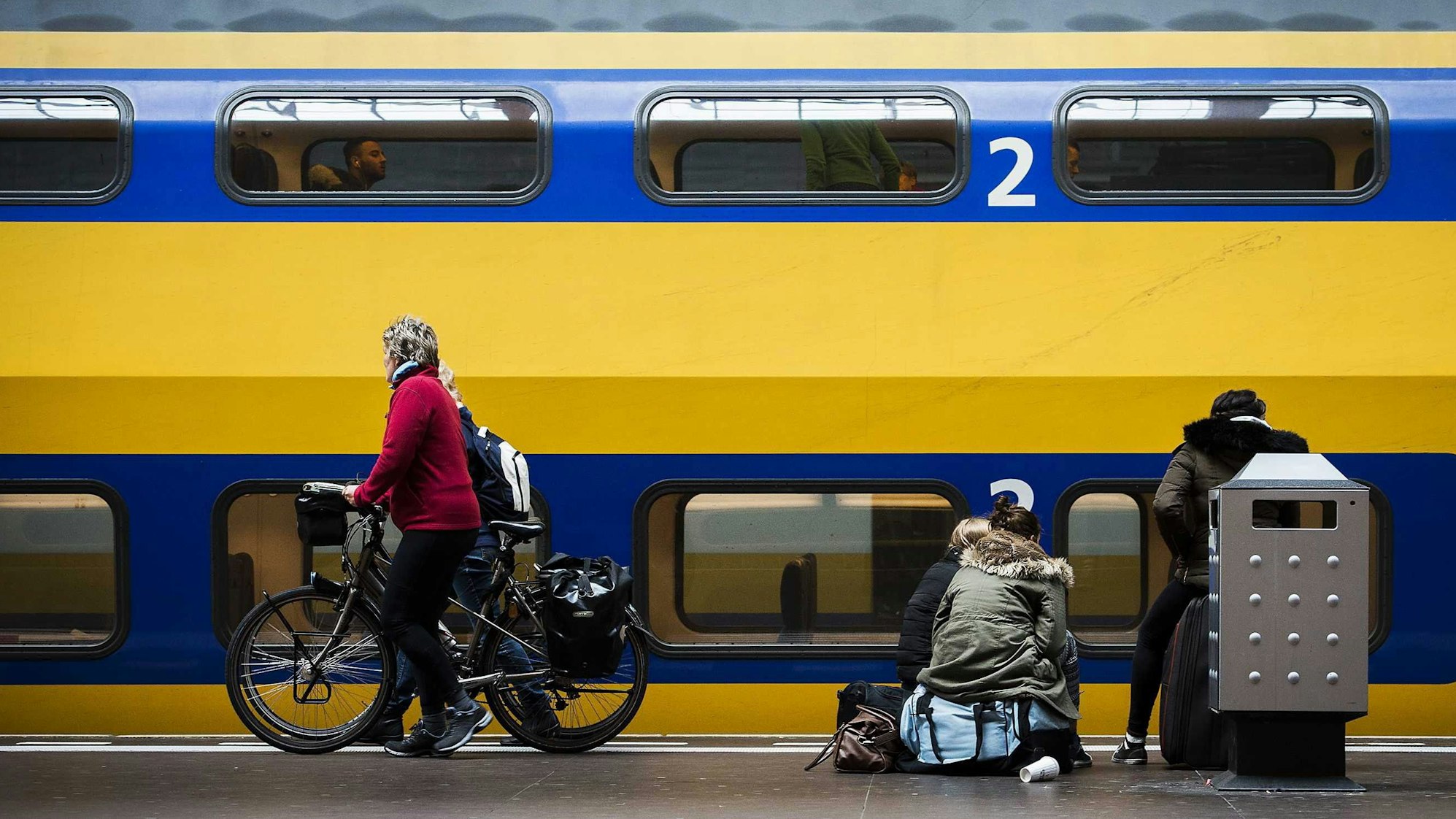 Ein Zug hält an einem Bahnhof in den Niederlanden. Vor dem Zug warte Passagiere, eine Frau in einer roten Jacke schiebt ein Fahrrad am Gleis entlang. (Archivbild)