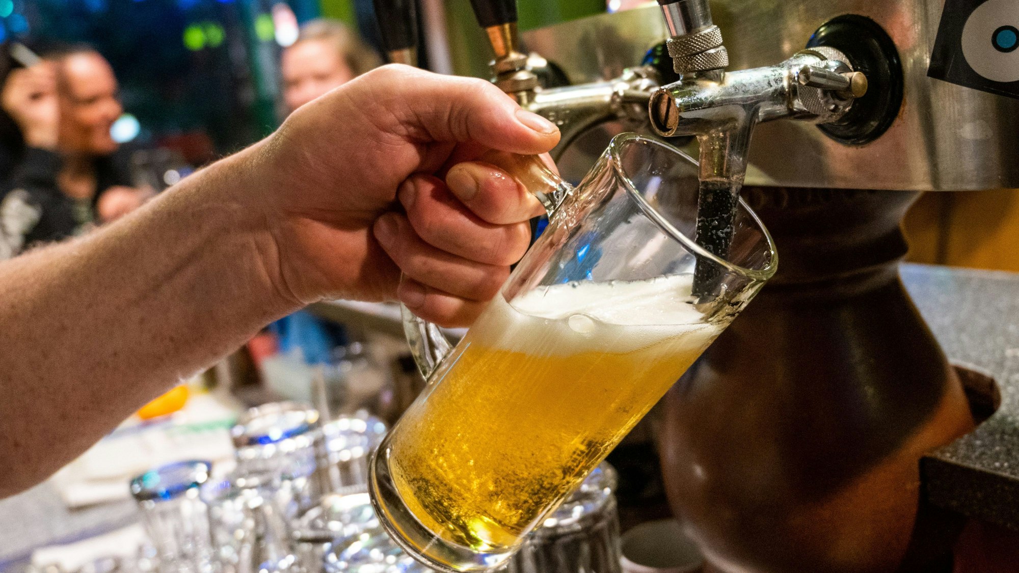 Eine Person zapft Bier. Im Hintergrund sitzen weitere Personen.
