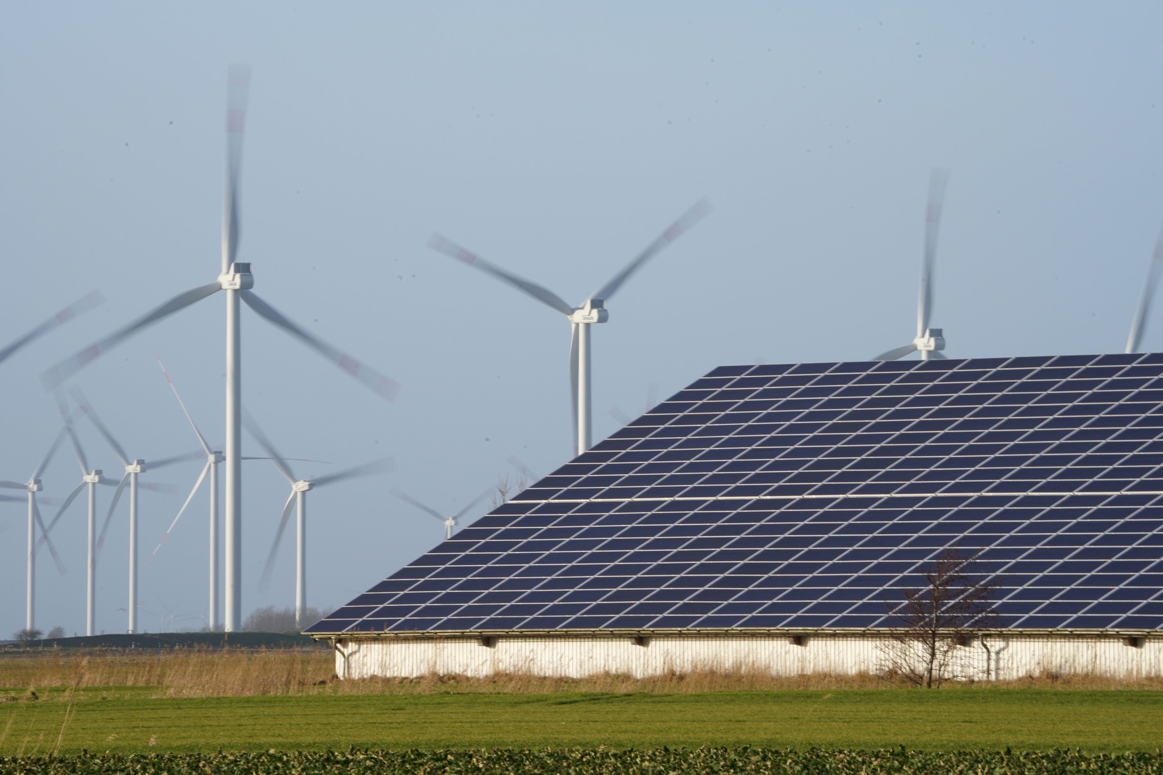 Windenergieanlagen stehen neben einer Halle mit Photovoltaik-Anlagen auf dem Dach.