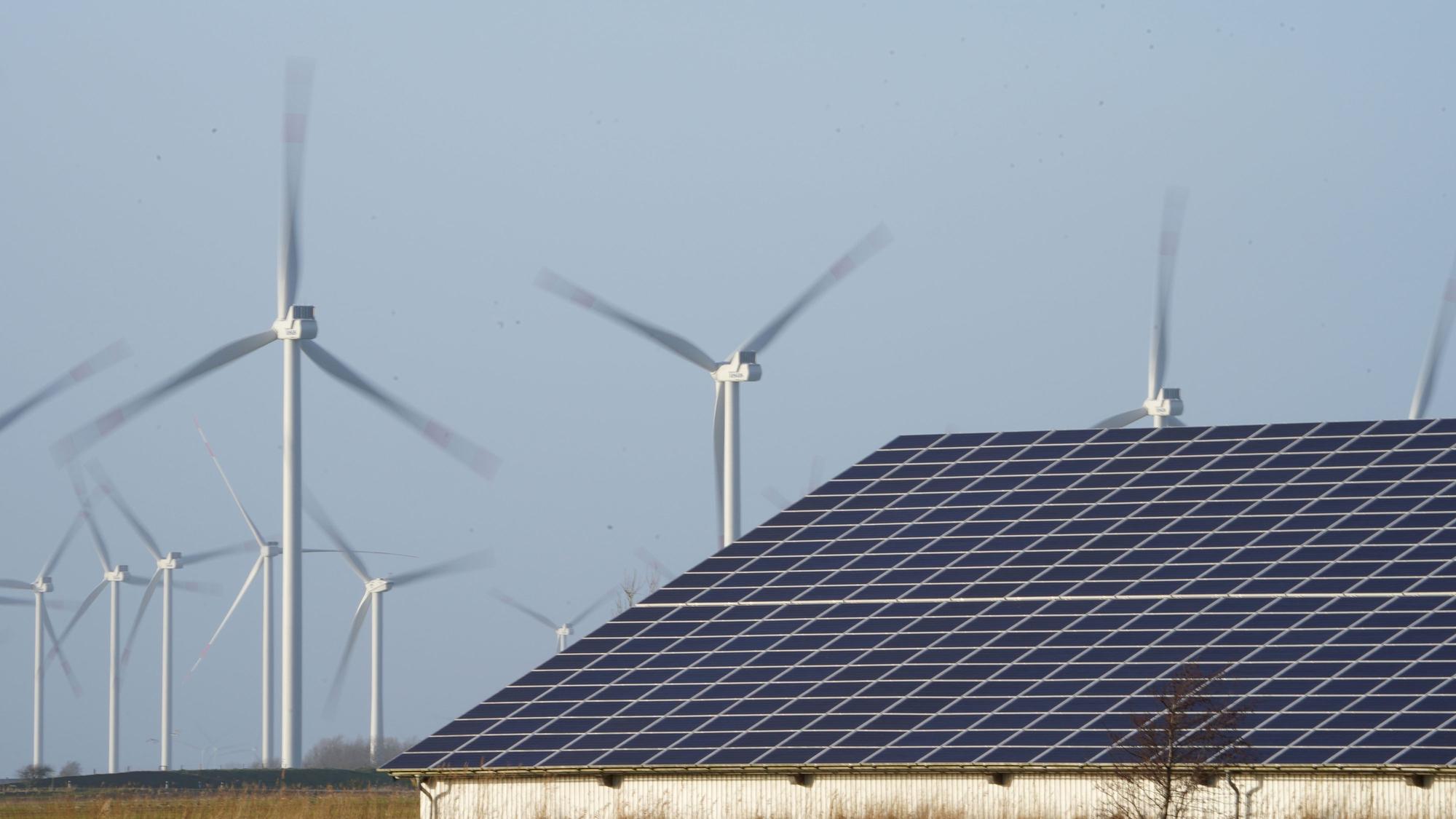 Windenergieanlagen stehen neben einer Halle mit Photovoltaik-Anlagen auf dem Dach.