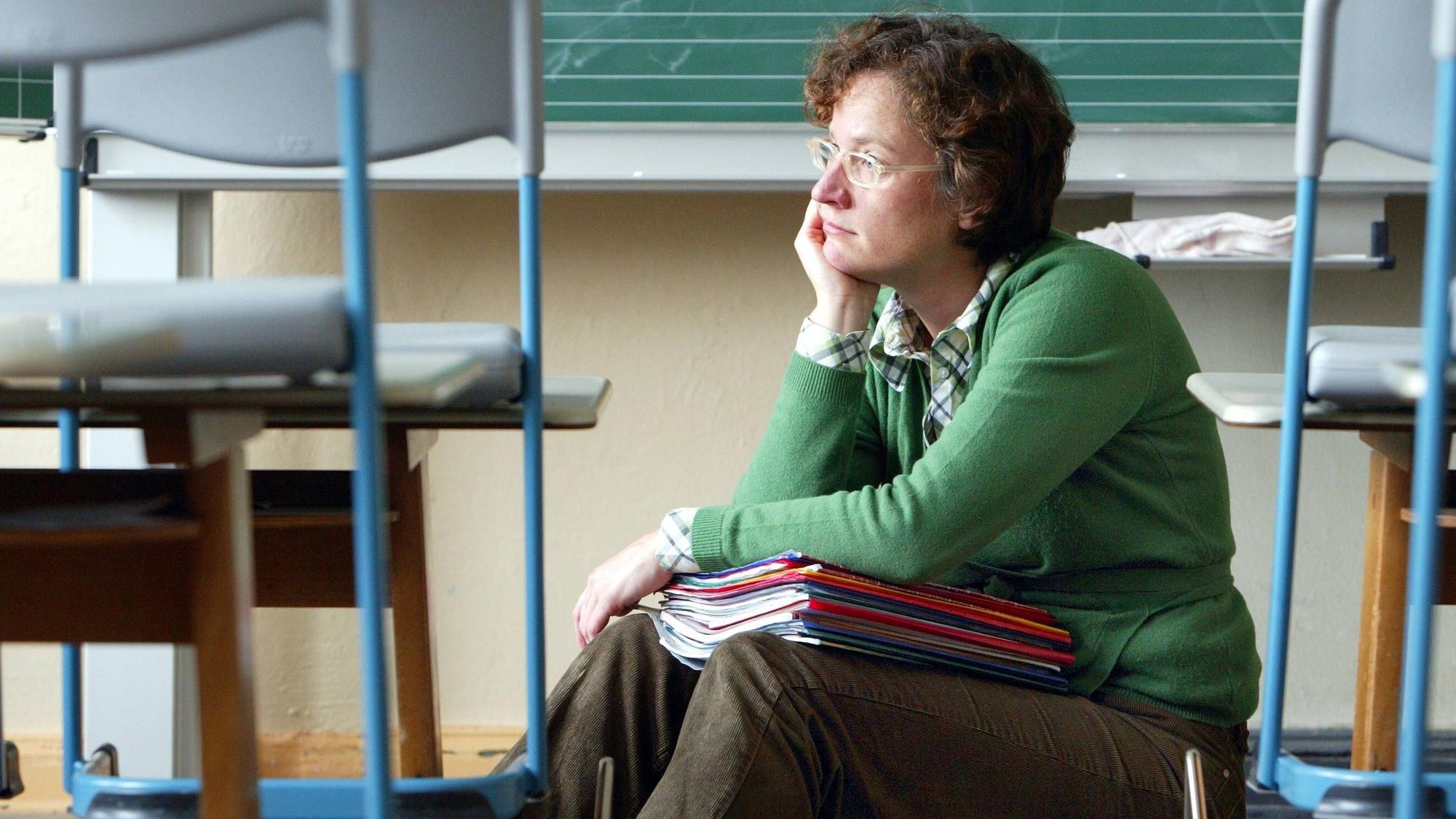 Eine Lehrerin sitzt alleine in einem Klassenraum mit einem Stapel Hefte auf dem Schoß.