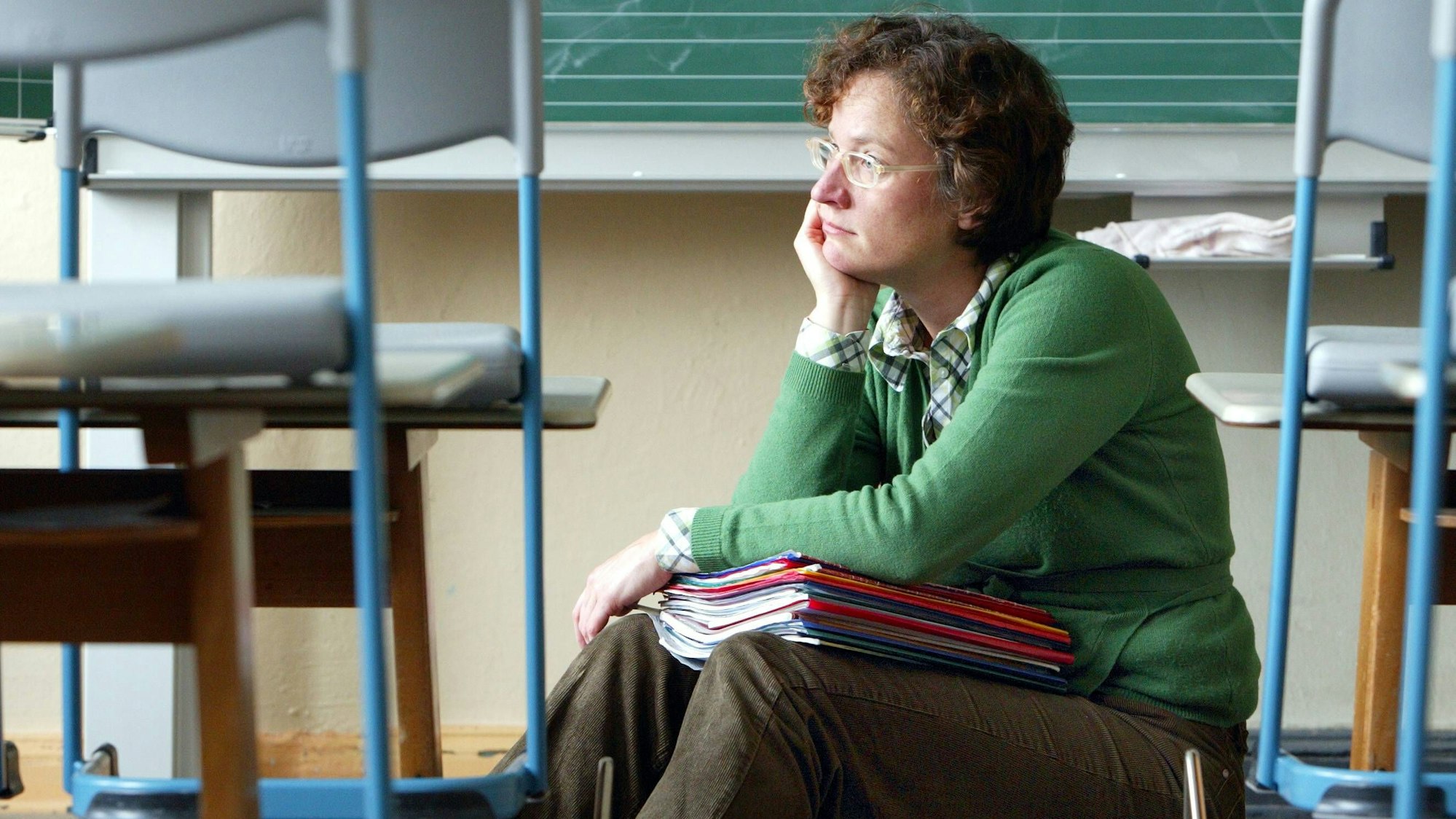 Eine Lehrerin sitzt alleine in einem Klassenraum mit einem Stapel Hefte auf dem Schoß.