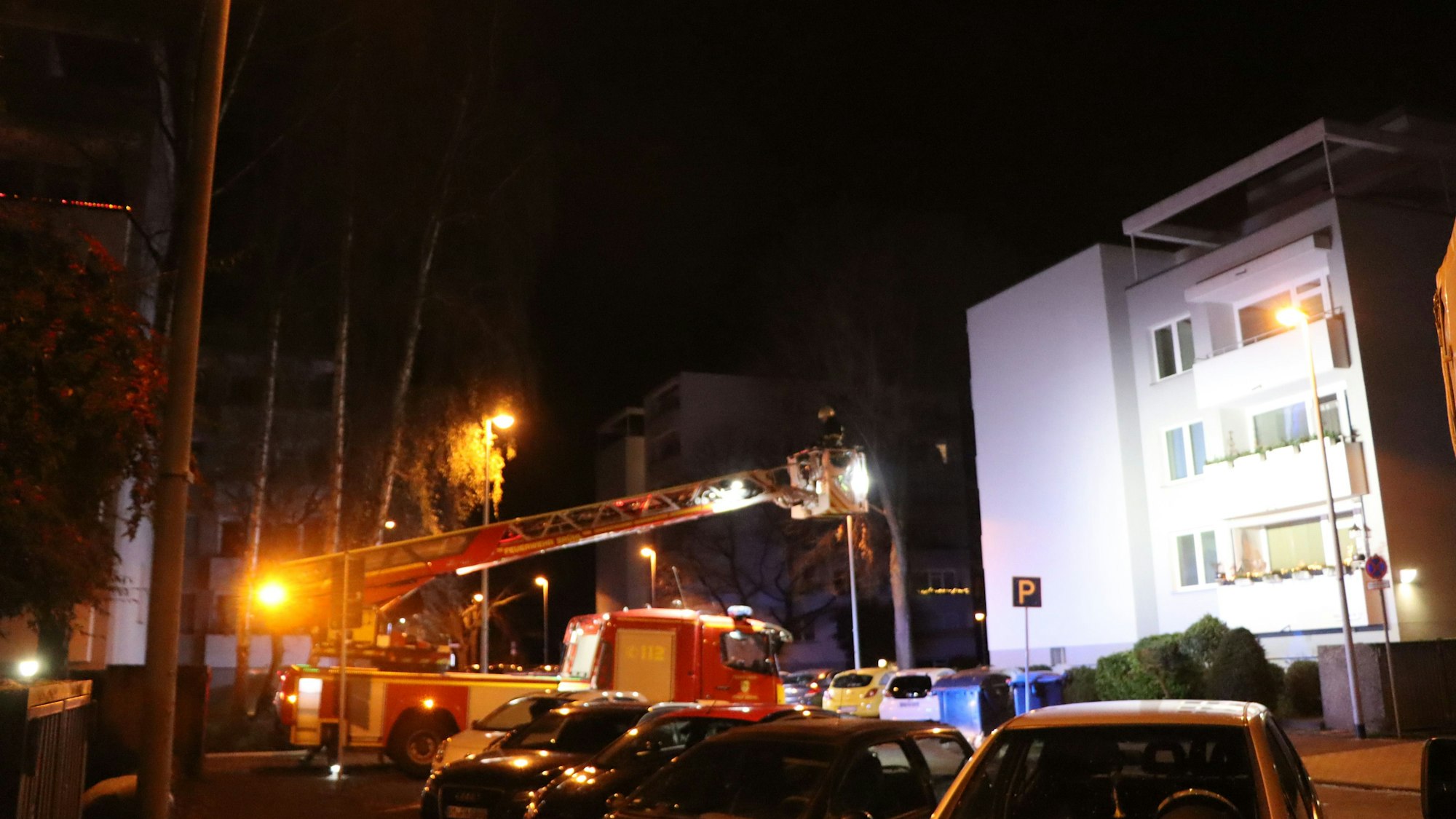 Die Feuerwehr dringt mit einer Drehleiter zum brennenden Mehrfamilienhaus in Brühl vor. Die weiße Fassade ist in der Dunkelheit angestrahlt.