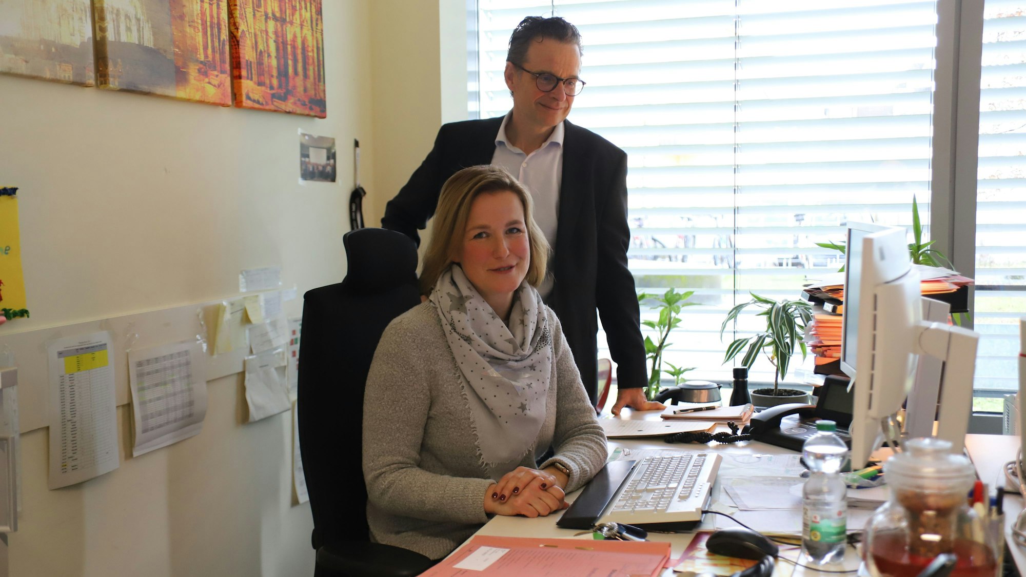 An einem Schreibtisch mit Aktenmappe, Tastatur und Computer-Monitor sitzt eine blonde Frau mit Pullover und Halstuch. Neben ihr steht ein Mann mit Brille, weißem Hemd und schwarzem Jackett.