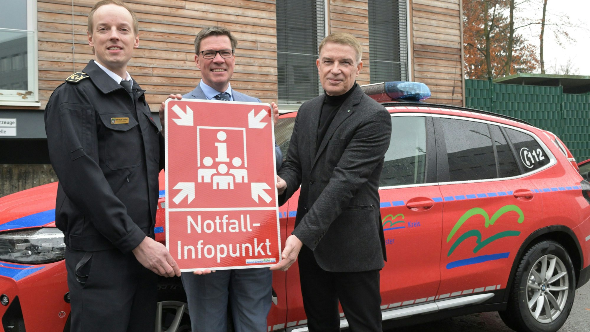 Kreisbrandmeister Martin Müller- Saidowski, Landrat Stephan Santelmann und Kreisdirektor Dr. Erik Werdel stehen vor der Kreisleitstelle und halten ein rotes NIP-Infoschild hoch.
