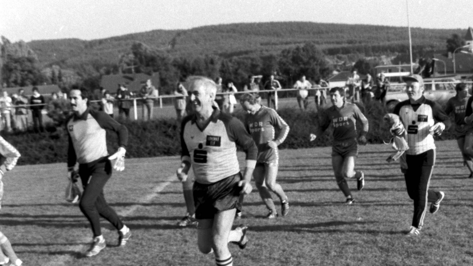 Die Mannschaften des WDR und der Linden-Elf laufen in zwei Reihen nebeneinander im Kaller Grenzlandstadion ein. Die Kreis-Kicker werden von Werner Schumacher (Vordergrund) angeführt.