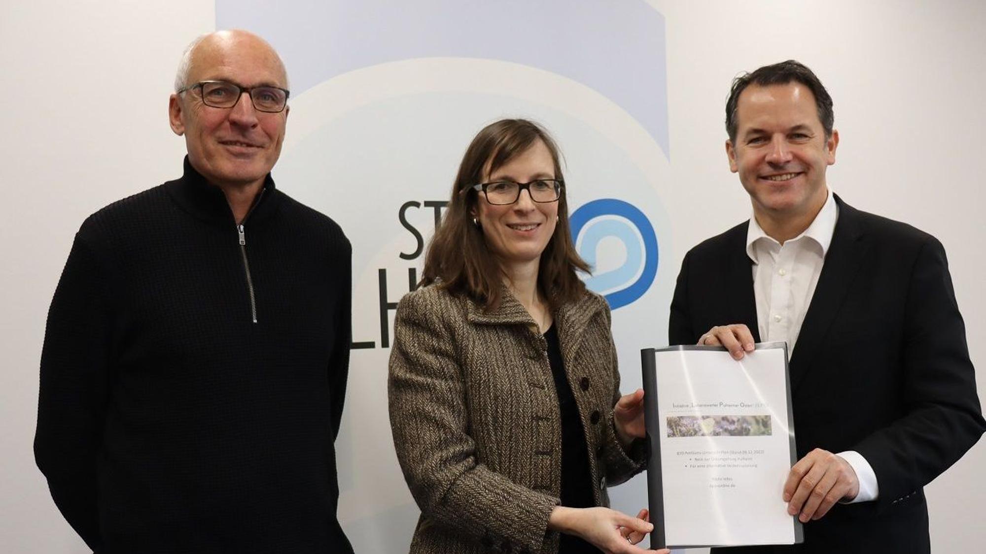 Frau steht zwischen zwei Männern und übergibt einem von ihnen eine Mappe mit Dokumenten