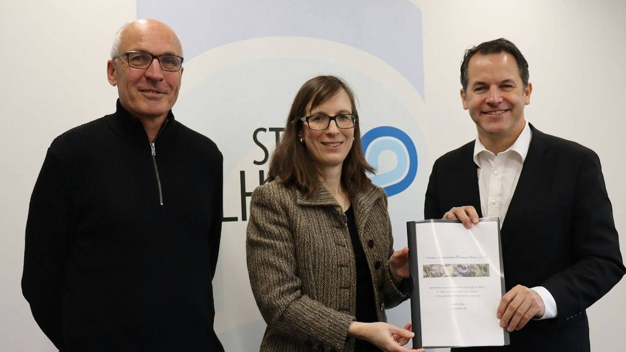 Frau steht zwischen zwei Männern und übergibt einem von ihnen eine Mappe mit Dokumenten