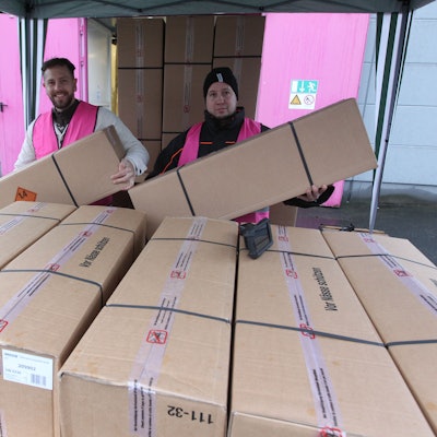 Zwei Männer in pinkfarbenen Weco-Westen heben zwei lange Pappkartons in die Höhe, vor ihnen steht ein hoher Stapel weiterer Kartons.