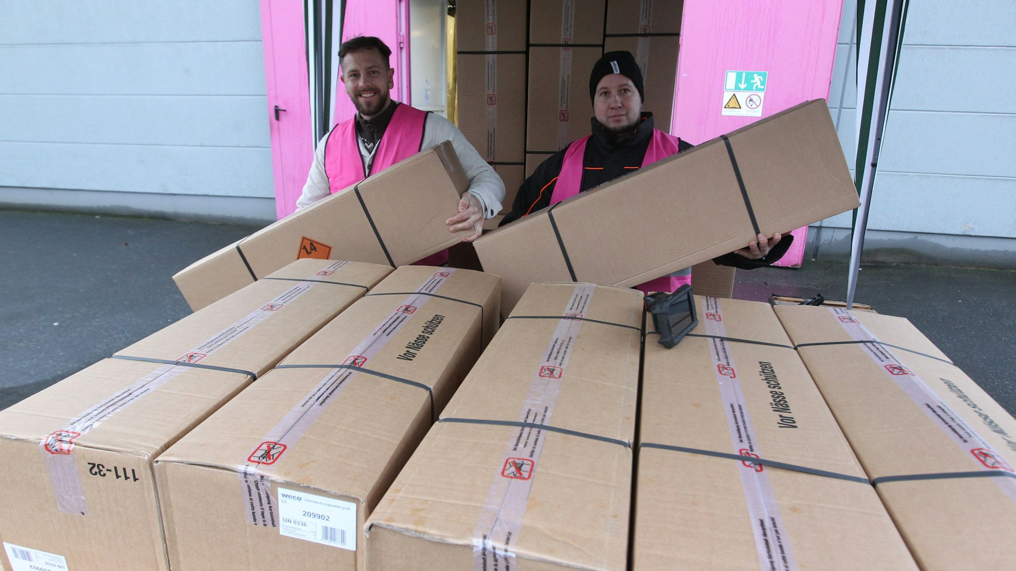 Zwei Männer in pinkfarbenen Weco-Westen heben zwei lange Pappkartons in die Höhe, vor ihnen steht ein hoher Stapel weiterer Kartons.