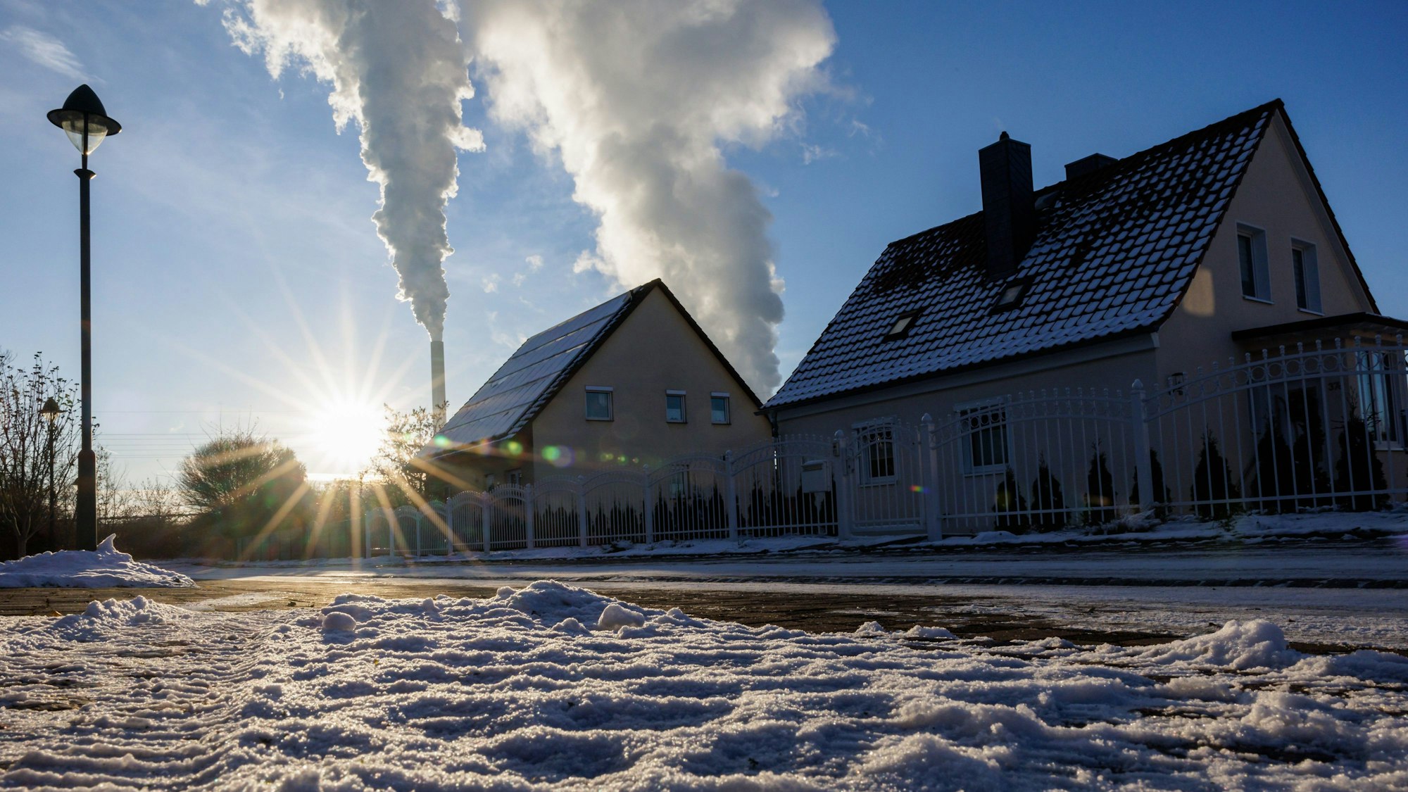Zwei Wohnhäuser im sonnenbeschienenen Schnee sind zu sehen. Dahinter steigen Rauchwolken aus hohen Schornsteinen in den blauen Himmel.