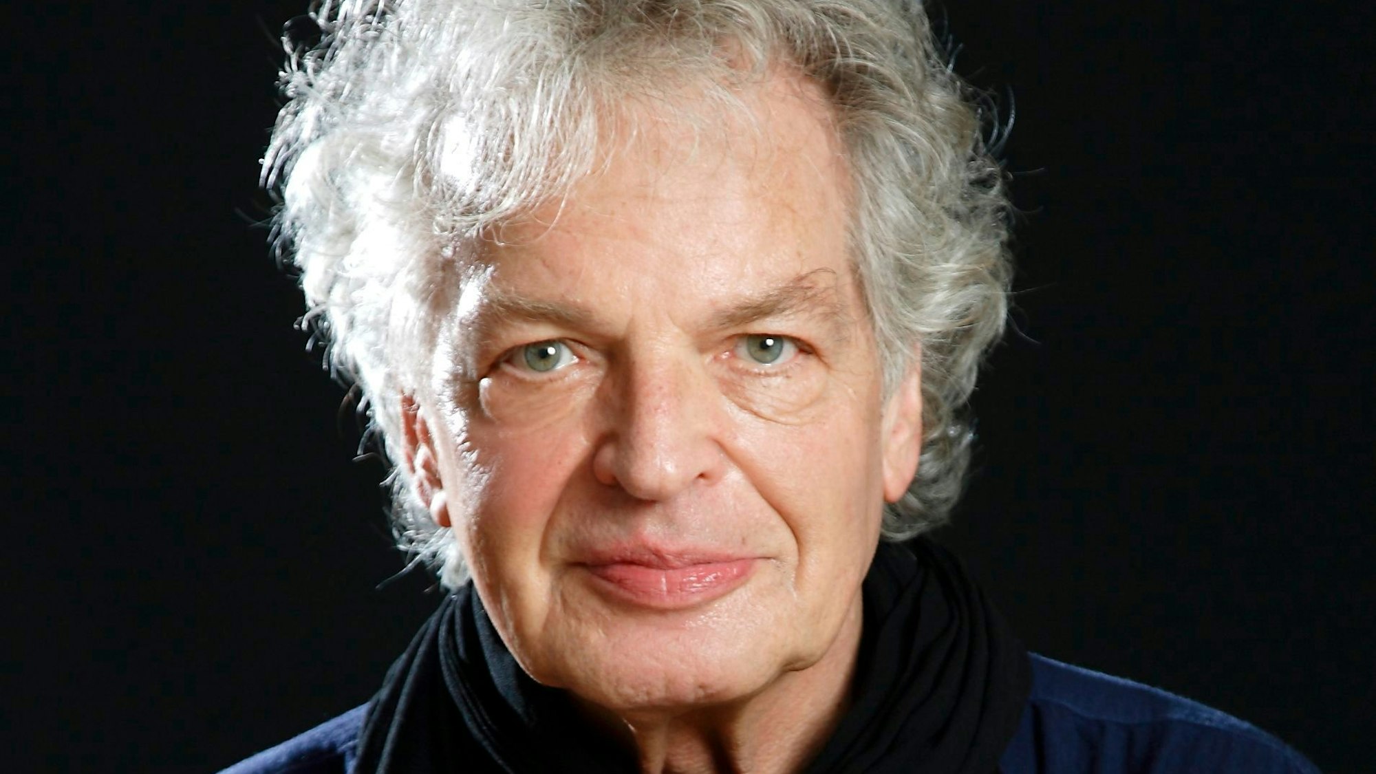 Ein Portrait des Jazz-Weltstars Joachim Kühn. Er trägt ein blaues Oberteil, einen schwarzen Schal und blickt mit sanften Gesichtszügen in die Kamera.