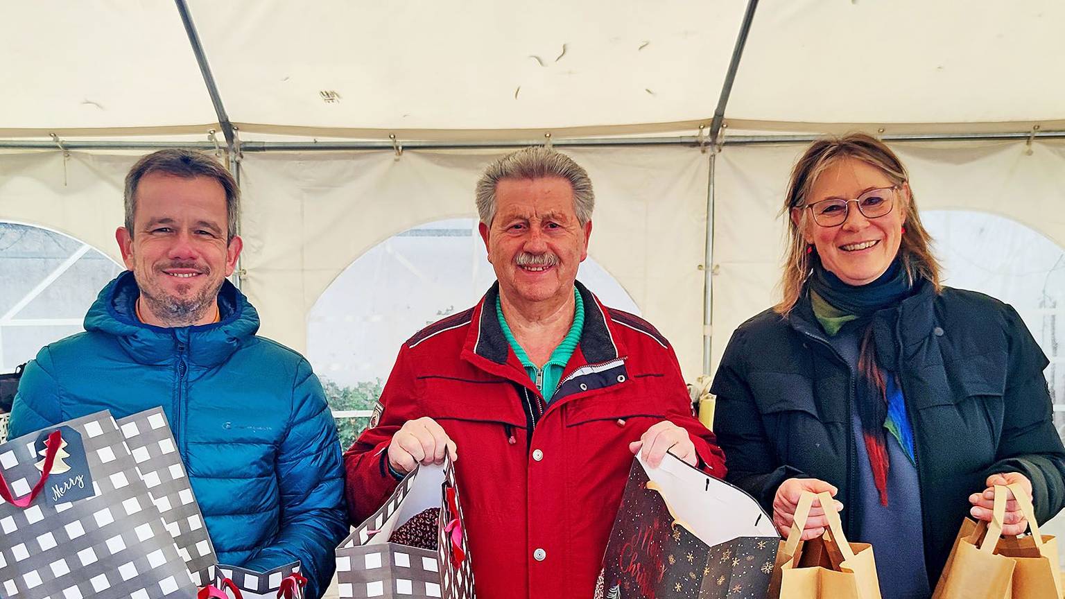 Martin Jost, Hubert Hamacher und Maria Surges-Brilon stehen unter einem Zelt und halten Weihnachtstüten in den Händen.