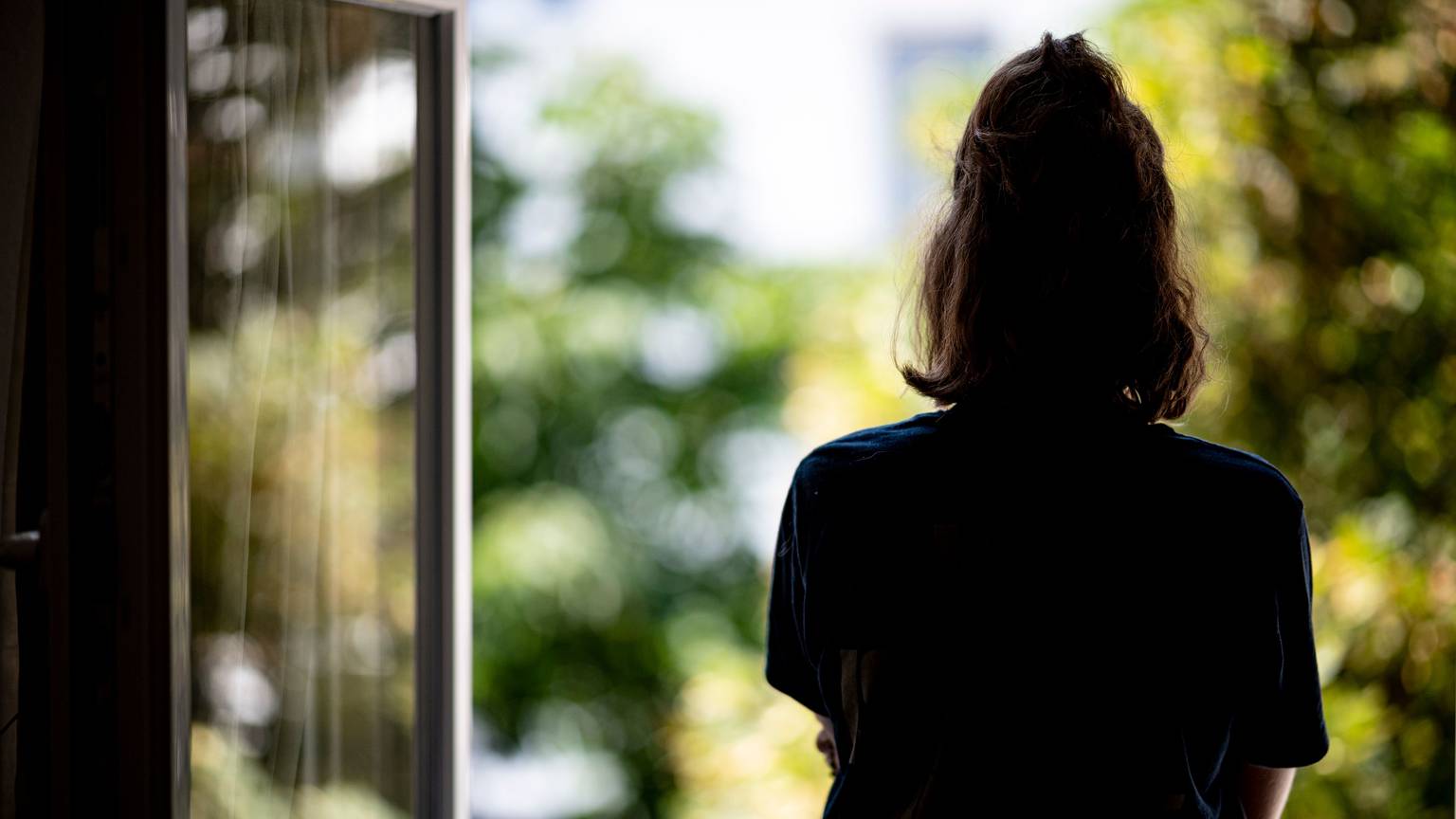 Eine Frau steht in ihrer Wohnung an einem geöffneten Fenster und blickt hinaus.