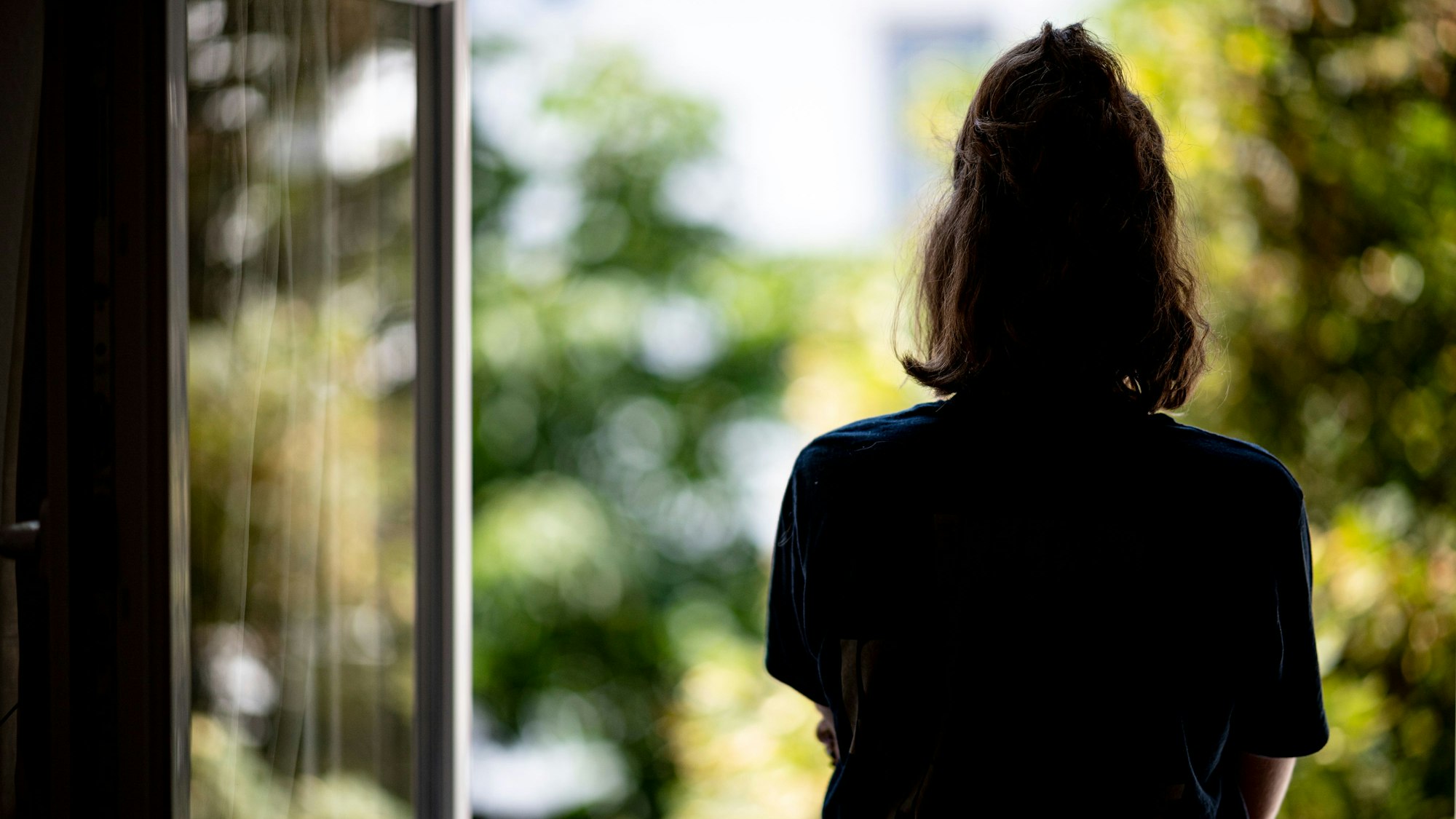 Eine Frau steht in ihrer Wohnung an einem geöffneten Fenster und blickt hinaus.
