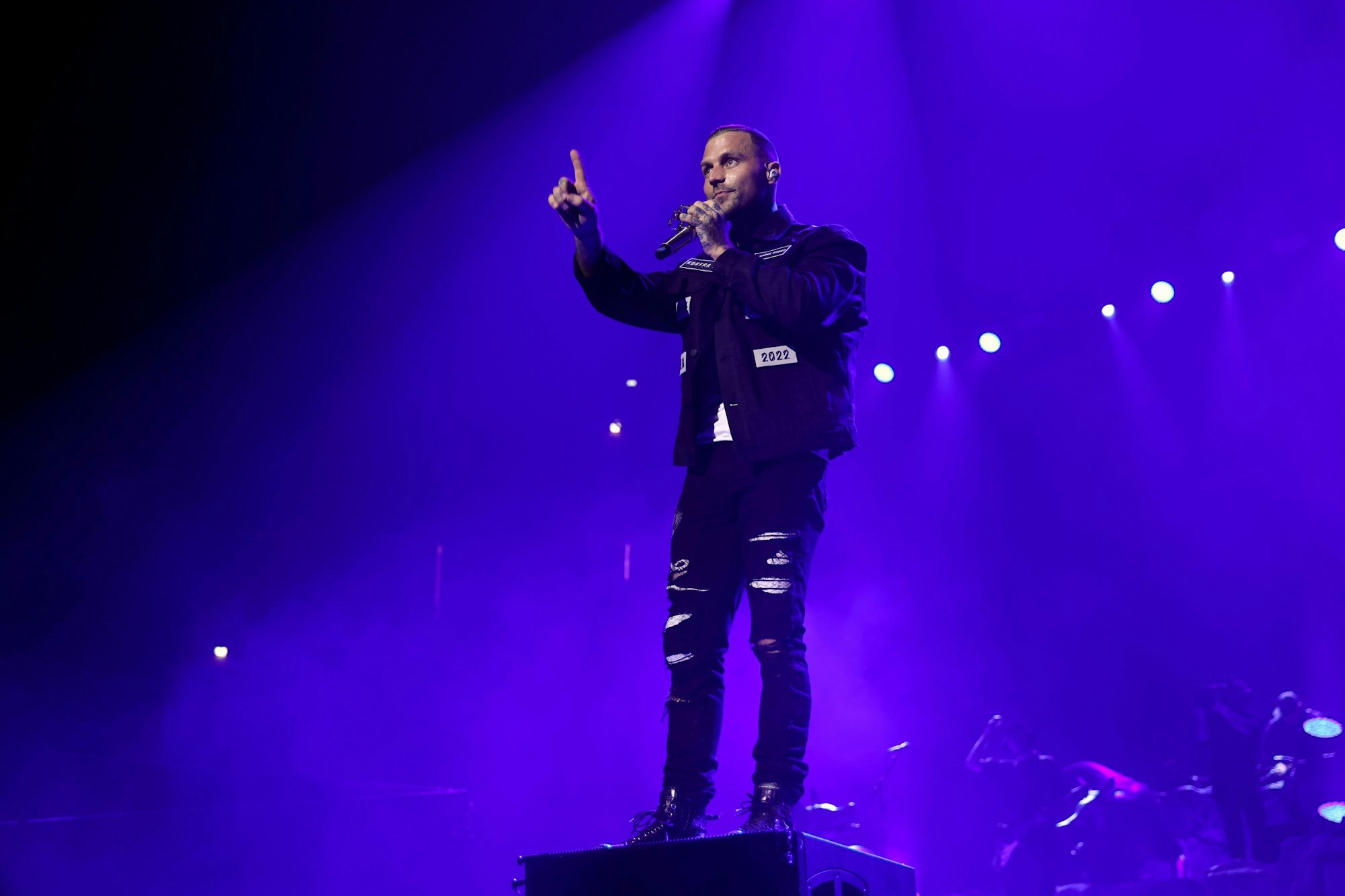 Der Künstler steht auf einem Podest und streckt den rechten Zeigefinger am ausgestreckten Arm in die Höhe. Er ist alleine auf dem Foto. Der Hintergrund ist dunkel und wird nur von violettem Scheinwerferlicht erhellt.