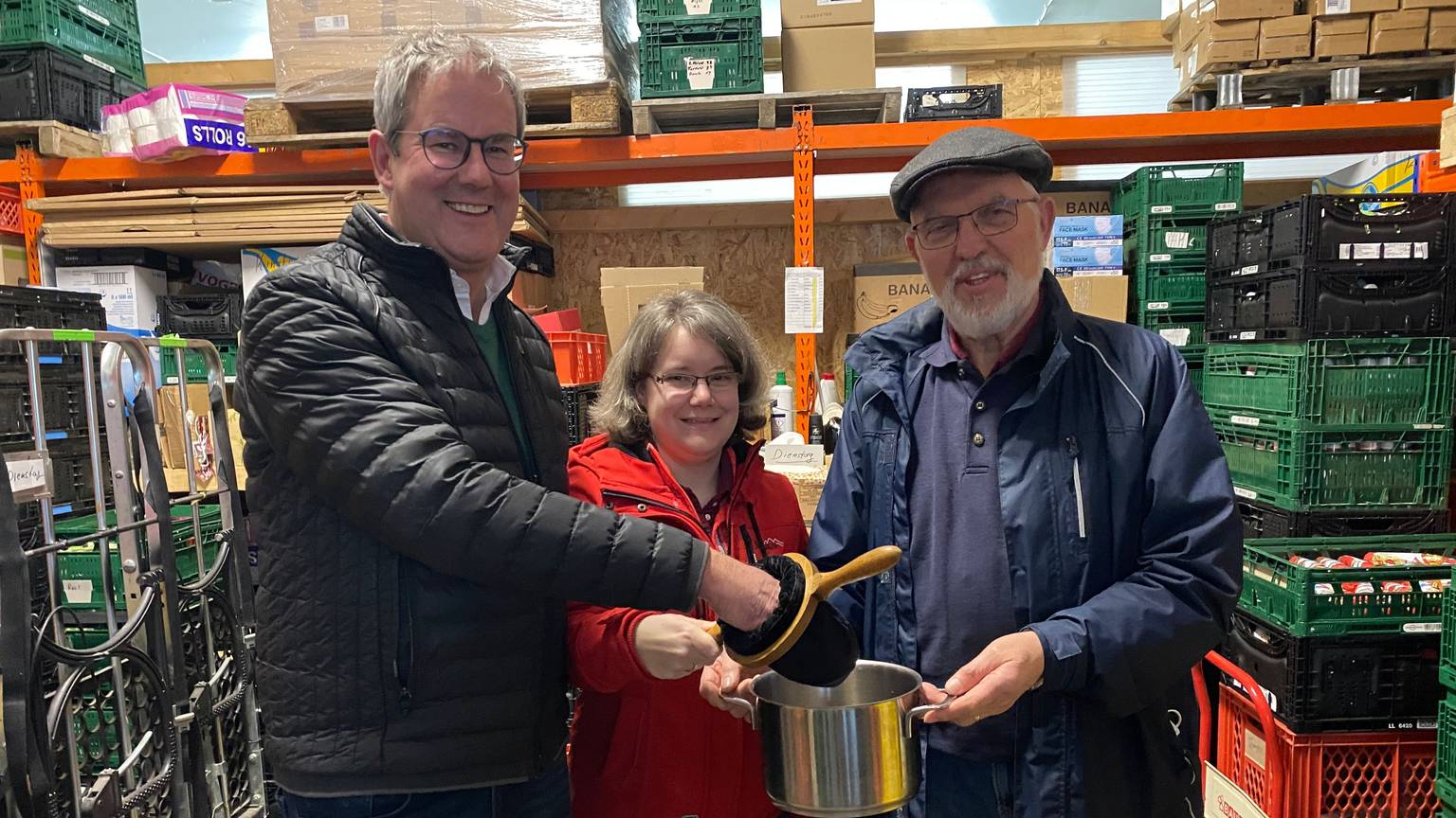 Pfarrer Frank Thönnes (v.l.), Tammy Peiter und Walter Feckinghaus stehen im Lager der Tafel Euskirchen und halten den Klingelbeutel in der Hand, der symbolisch in einem Topf entleert wird.