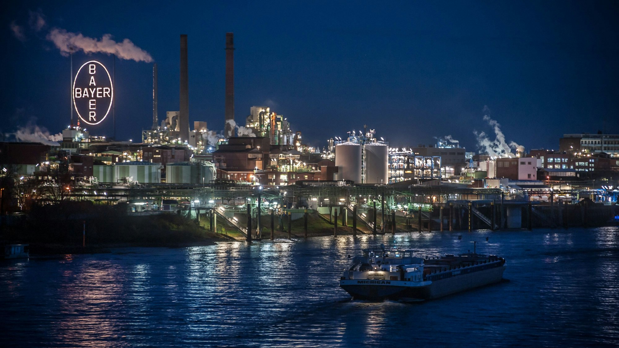 Vorne ist der Rhein bei Nacht zu sehen, dahinter erhebt sich der Chempark mit Anlagen, dampfenden Schornsteinen und dem leuchtenden Bayer-Kreuz.