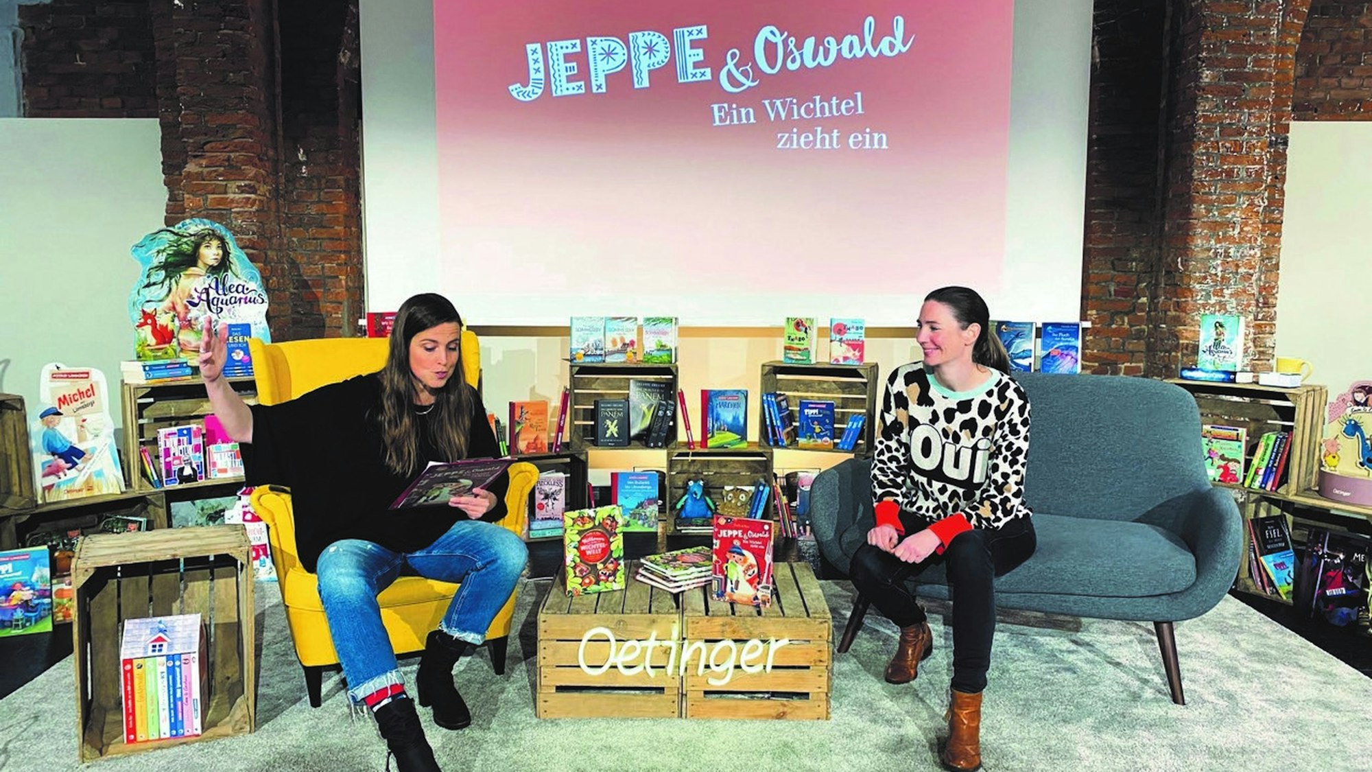 Eva Dax und Sabine Dully bei der Präsentation des Buches. Sie sitzen inmitten vieler Kinderbücher vor einer Leinwand.