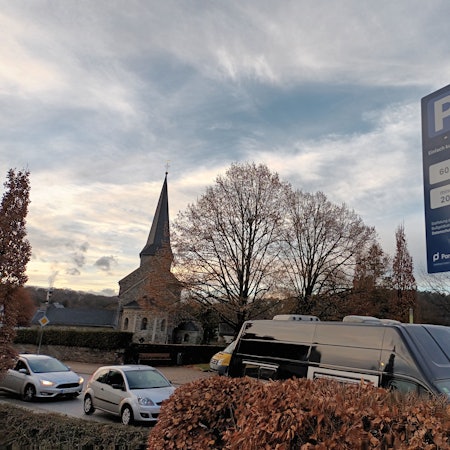 Ein kleines, blaues Schild auf einem Supermarkt-Parkplatz im Lohmarer Zentrum informiert über die Höchstdauer fürs Parken und das Verwarngeld von mindestens 20 Euro. Im Hintergrund ist die katholische Kirche zu sehen.