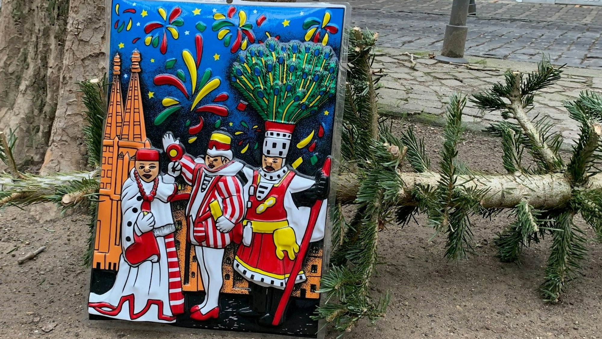 Auf der Weißenburgstraße im Agnesviertel lehnt ein Plastikbild mit dem Kölner Dreigestirn am 30. Dezember 2022 an den Resten eines Weihnachtsbaums.