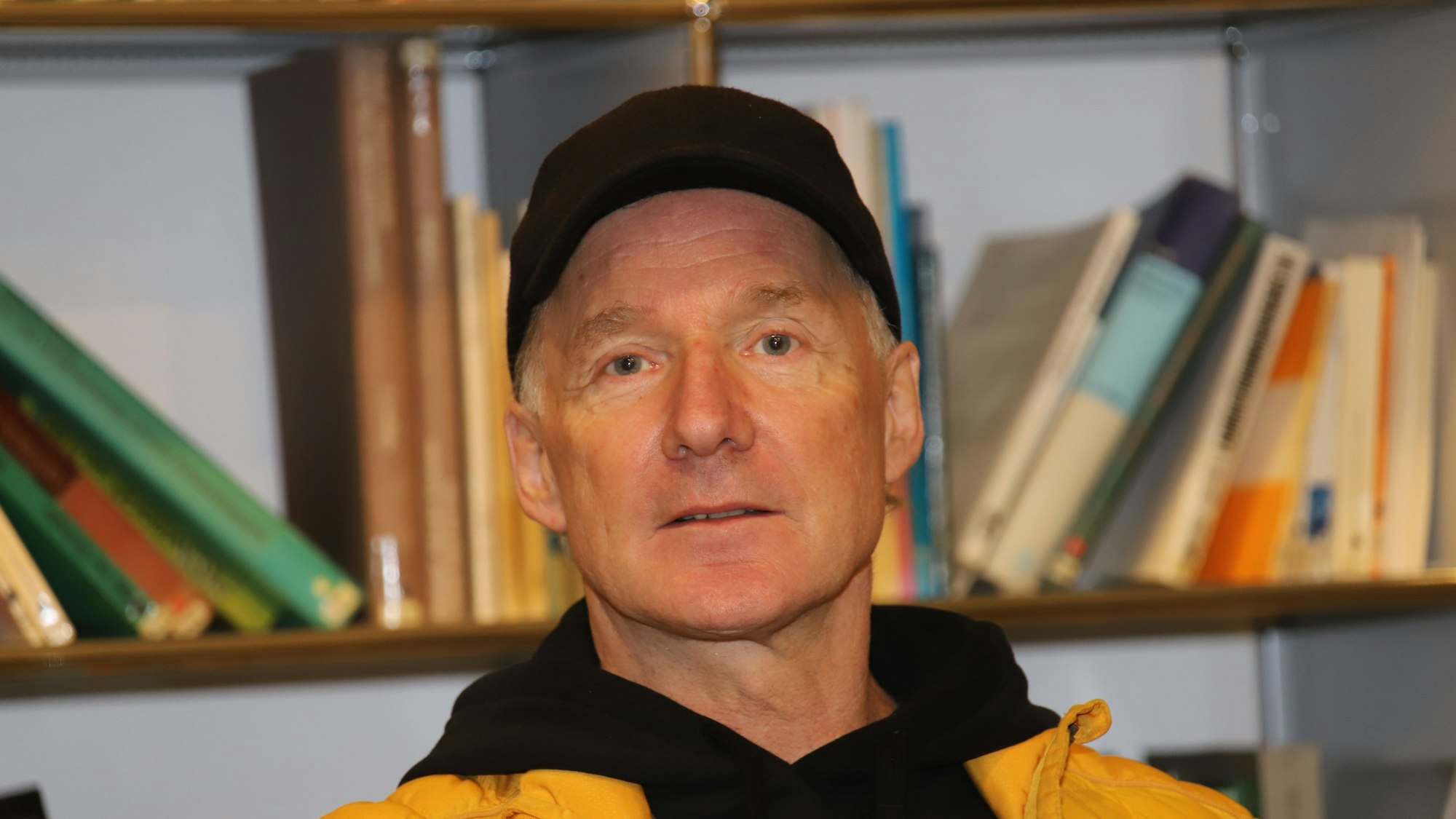 Ein Mann mit Käppi, Kapuzenpullover und Jacke steht vor einem Regal mit Büchern.