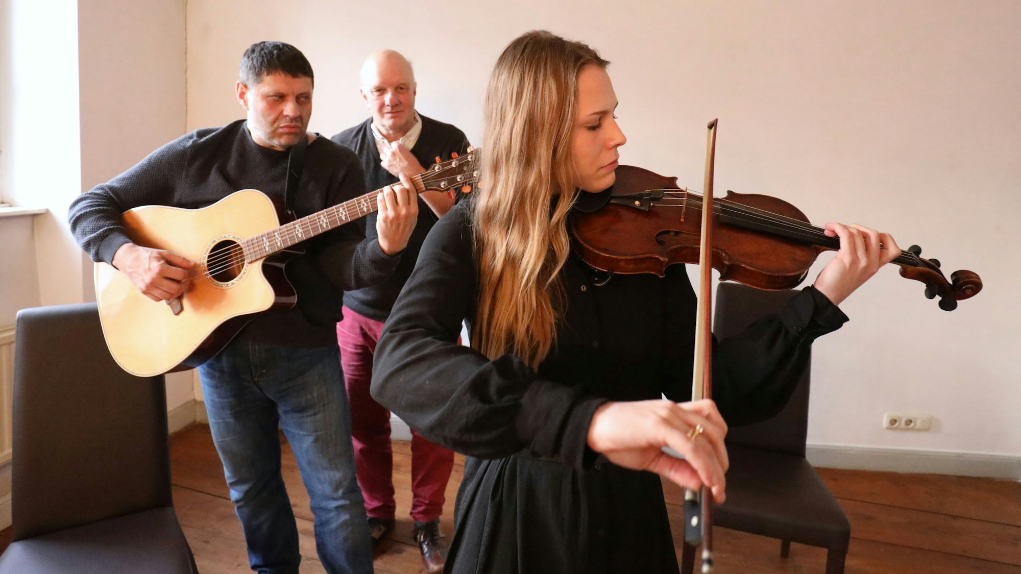 Eine junge Frau spielt Geige, hinter ihr spielt ein Mann die Gitarre. Volker Hartung, Leiter der Jungen Philharmonie Köln, steht hinter den beiden.