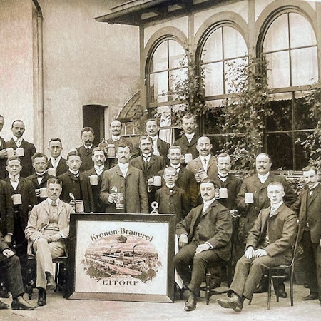 Das Bild zeigt eine Gruppe Männer mit Bierkrügen in der Hand. Sie posieren vor der Brauerei für ein Foto.