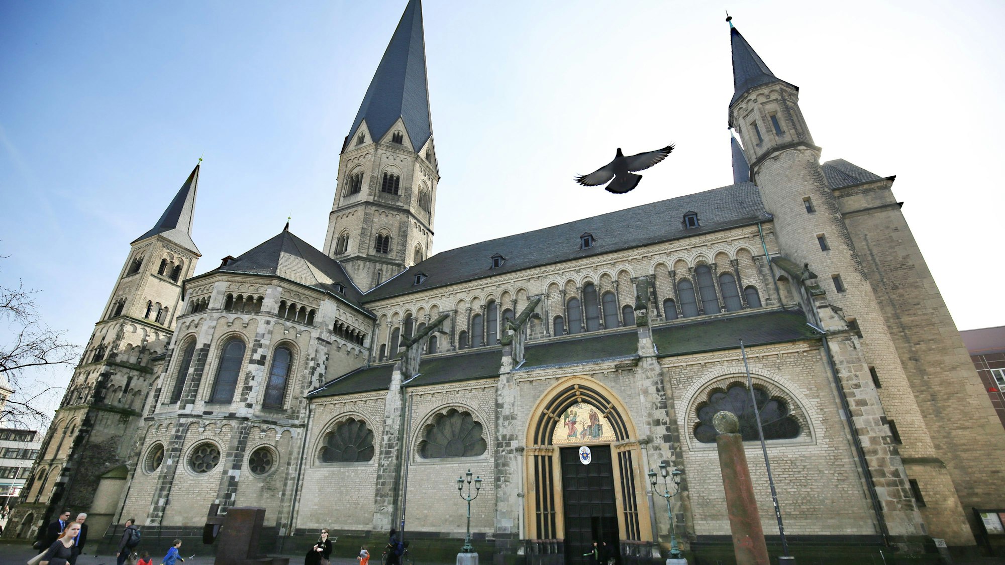 Vor einer alten Kirche mit mehreren Türmen und einem prächtigen Eingang laufen Menschen, eine Taube fliegt in die Höhe.