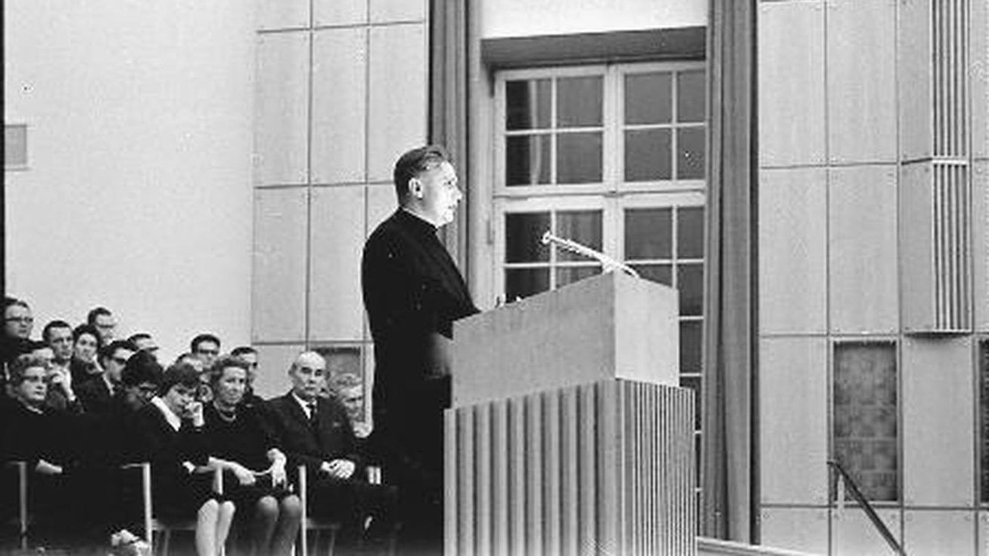 Ein altes Schwarz-Weiß-Foto zeigt einen schwarz gekleideten Mann an einem Rednerpult mit Mikrofon, hinter ihm sitzen mehrere Menschen.