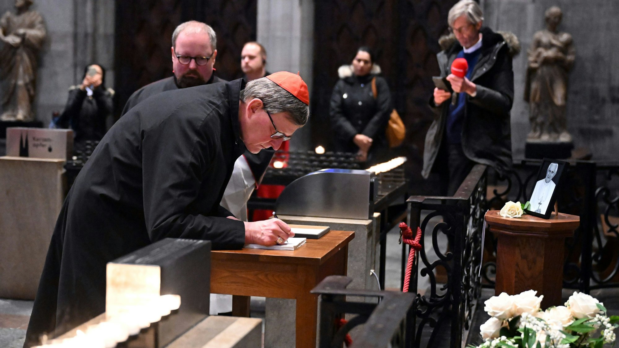 Kardinal Rainer Maria Woelki trägt sich in das Kondolenzbuch für den emeritierten Papst Benedikt XVI. im Kölner Dom ein. Vor dem Kondolenzbuch steht ein Bild des Papstes.