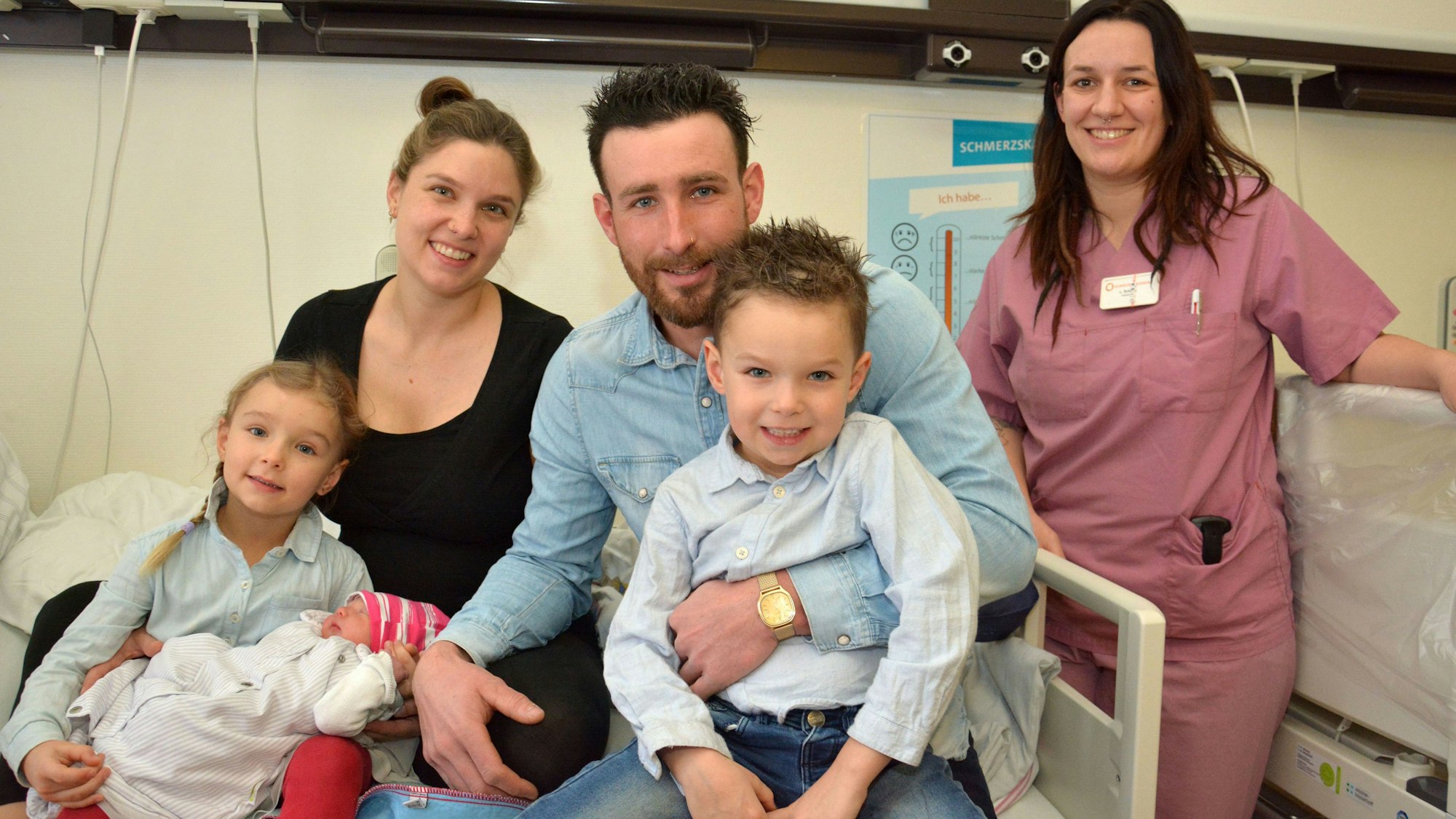 Zu sehen ist eine fünfköpfige Familie mit einem Neugeborenen und der Hebamme in einem Krankenhauszimmer.