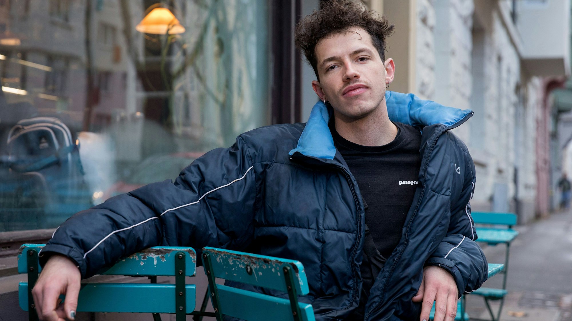 Paul Köninger sitzt auf einem Stuhl. Er trägt eine blaue Jacke und hat seinen rechten Arm auf der Armlehne eines zweiten Stuhls.