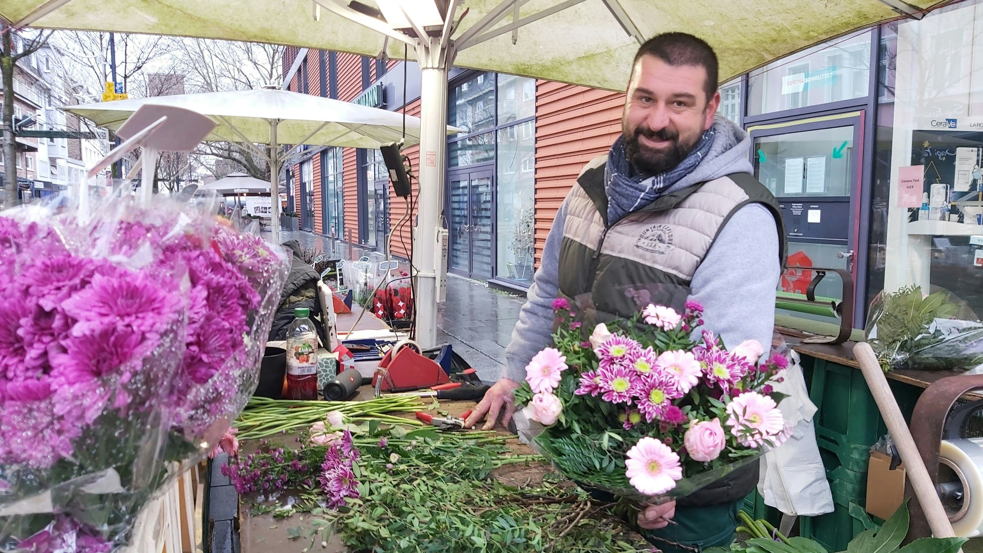 Ein Mann in Pulli und wärmender Weste präsentiert einen bunten Strauß an seinem Blumenstand.