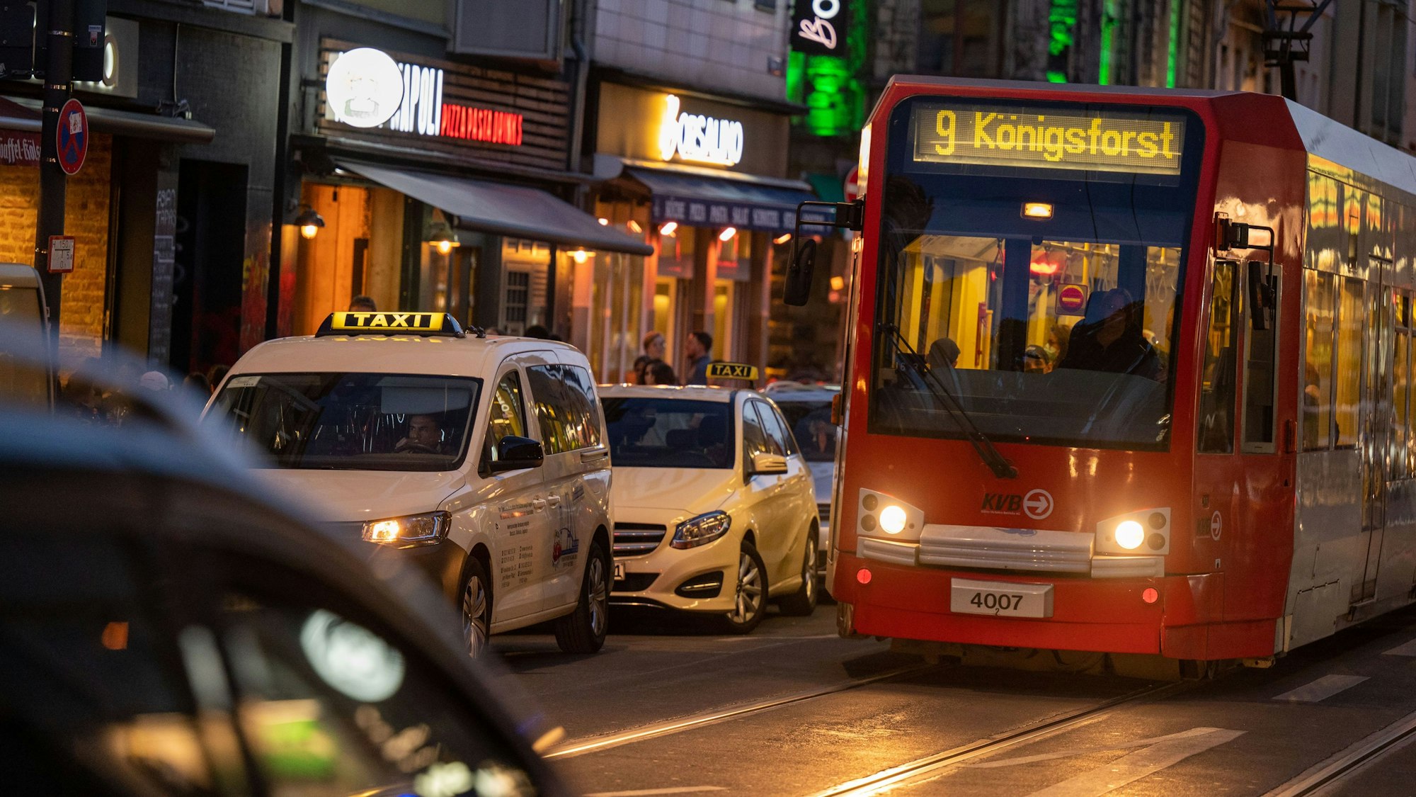 Eine Straßenbahn der Linie 9 fährt in der Dämmerung auf der Zülpicher Straße an geparkten Taxis vorbei.