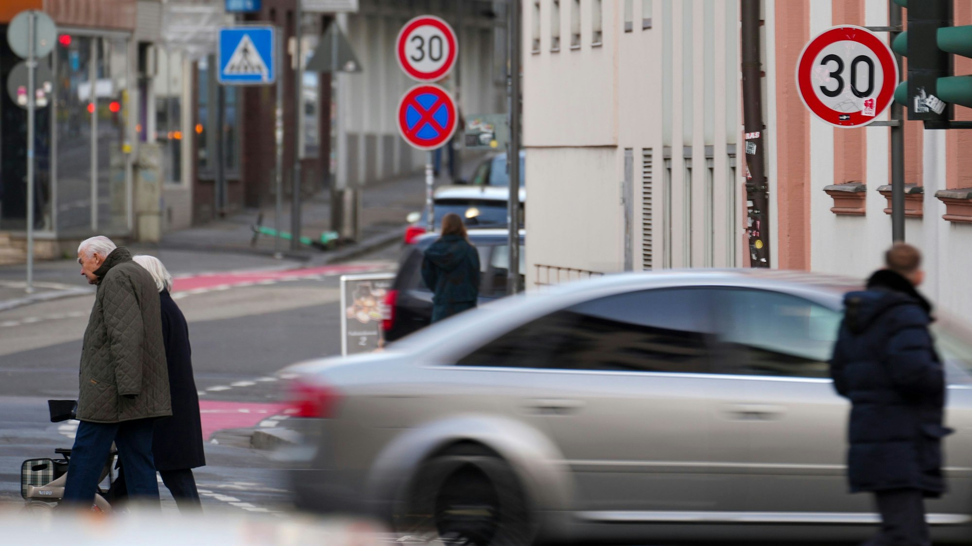 In der Innenstadt fährt ein Auto, Fußgänger überqueren die Straße. Um sie herum sind Ampeln und die Verkehrsschilder mahnen zu Tempo 30.