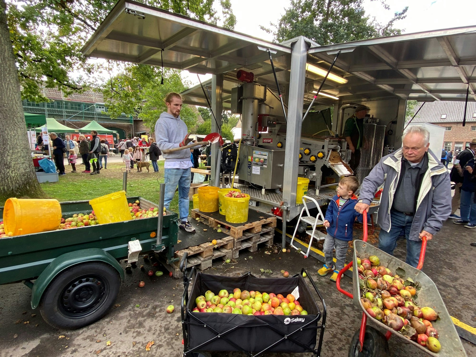 Eine Obstpresse presst viele Äpfel zu Saft – zwei Männer und ein Kind sind zu sehen.