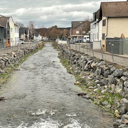 Die Erft fließt zwischen zwei Häuserreihen hindurch. Begrenzt wird sie durch wenige Steine und einen Bauzaun.
