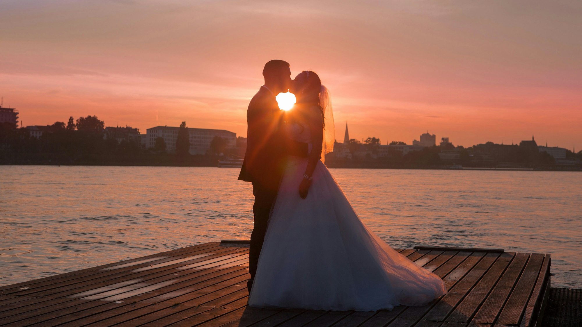 Ein Hochzeitspaar steht im Sonnenuntergang auf einem Steg vor einem Fluss.