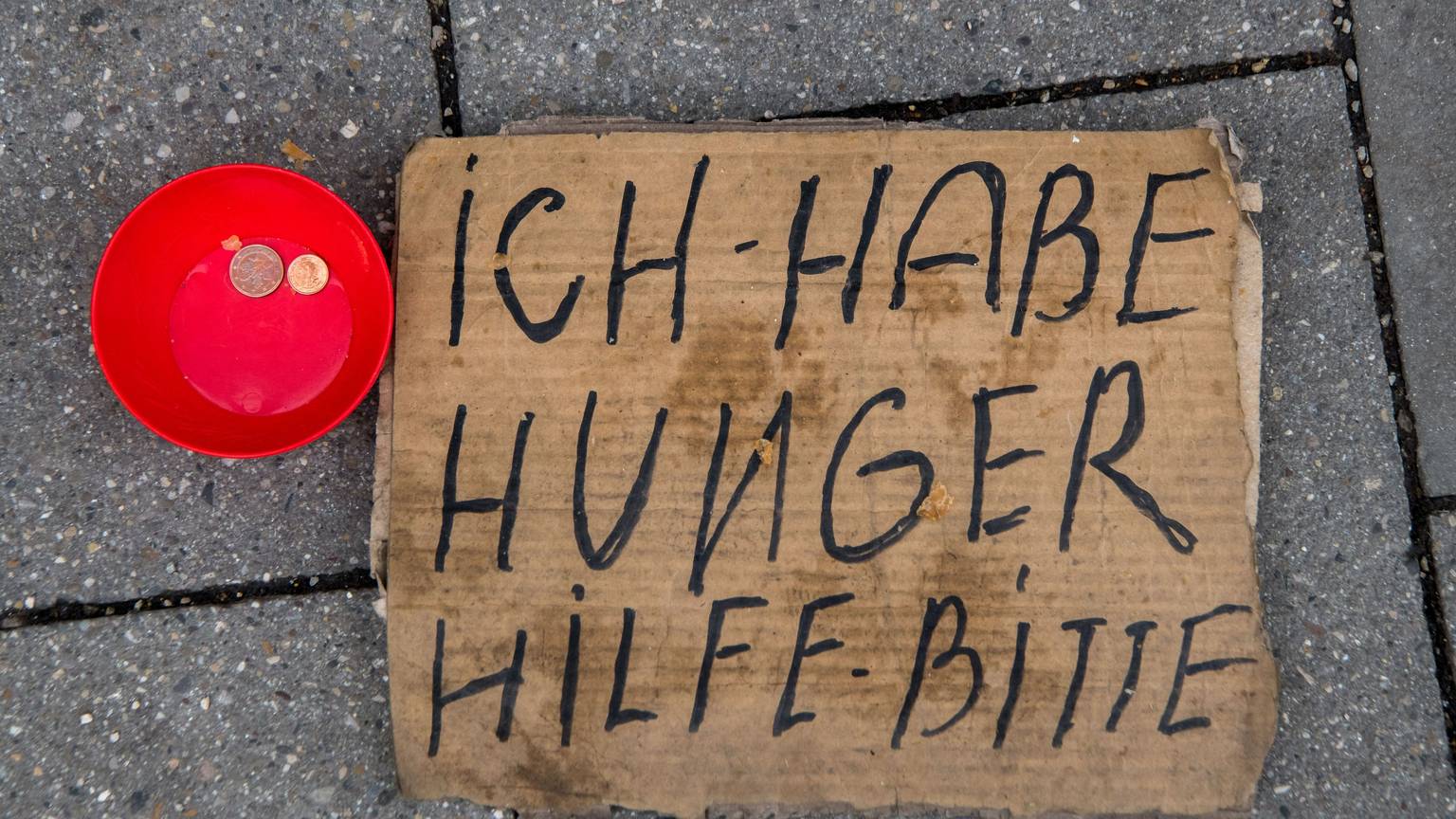 Ein dreckiges Pappschild mit der Aufschrift Ich habe hunger Hilf - bitte, liegt auf der Straße. Daneben steht eine rote kleine Schüssel mit zwei Münzen drinnen.
