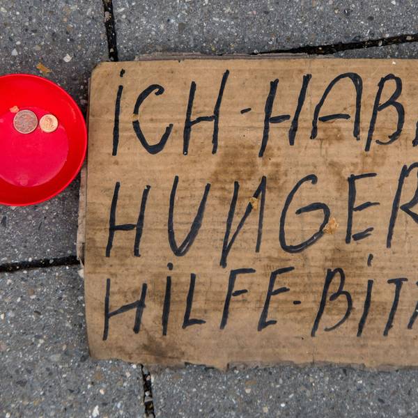 Ein dreckiges Pappschild mit der Aufschrift Ich habe hunger Hilf - bitte, liegt auf der Straße. Daneben steht eine rote kleine Schüssel mit zwei Münzen drinnen.