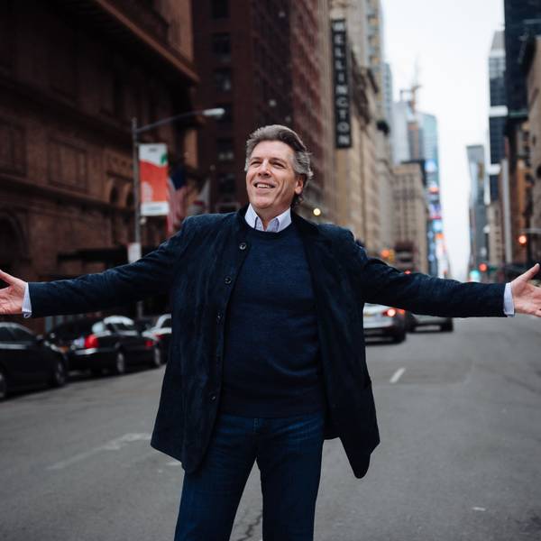 Thomas Hampson steht auf einer Straße und breitet die Arme aus. Hinter ihm Autos, Ampeln und Wolkenkratzer.