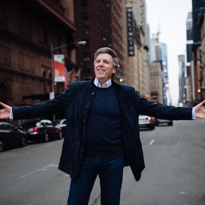 Thomas Hampson steht auf einer Straße und breitet die Arme aus. Hinter ihm Autos, Ampeln und Wolkenkratzer.