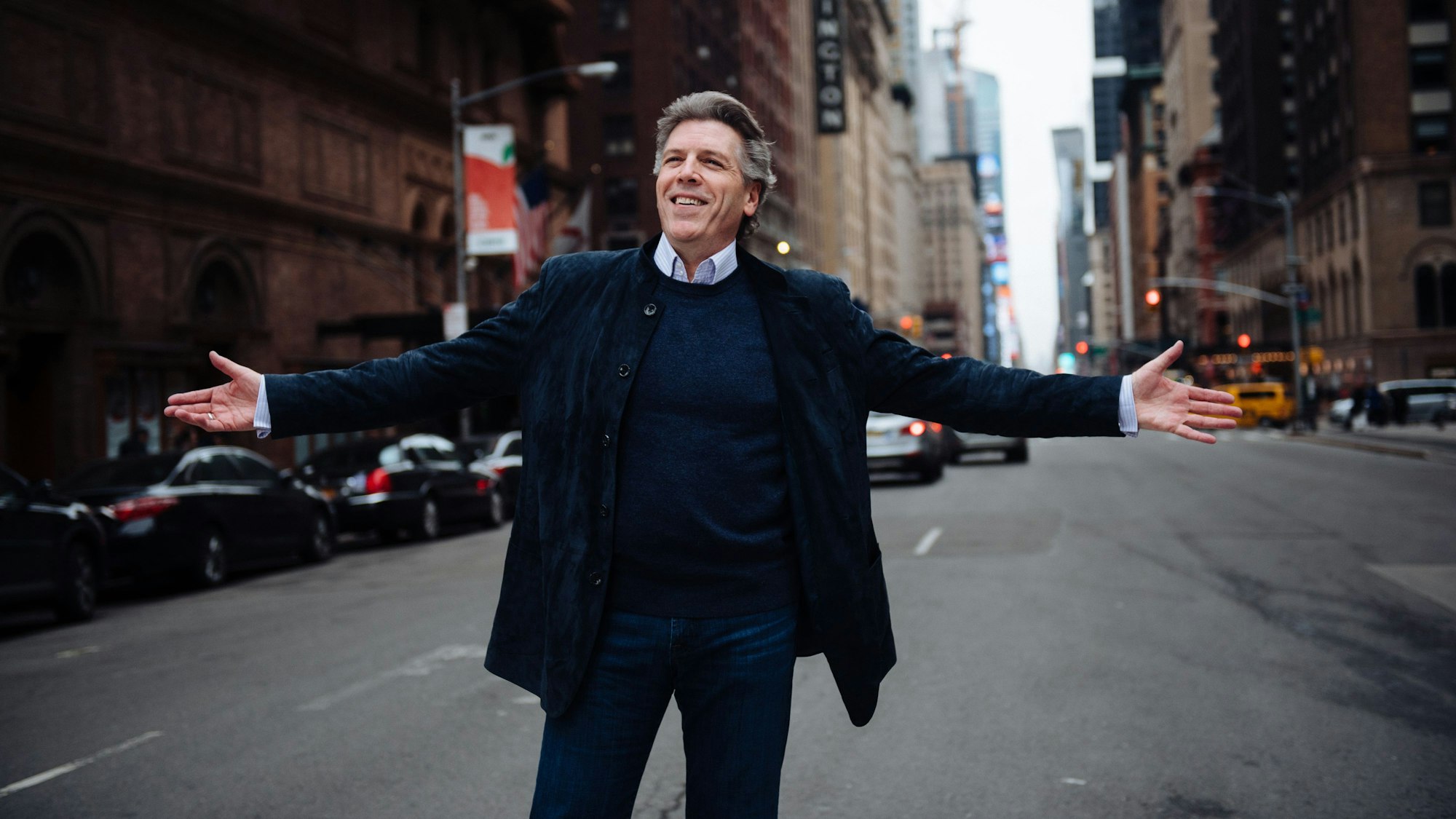 Thomas Hampson steht auf einer Straße und breitet die Arme aus. Hinter ihm Autos, Ampeln und Wolkenkratzer.