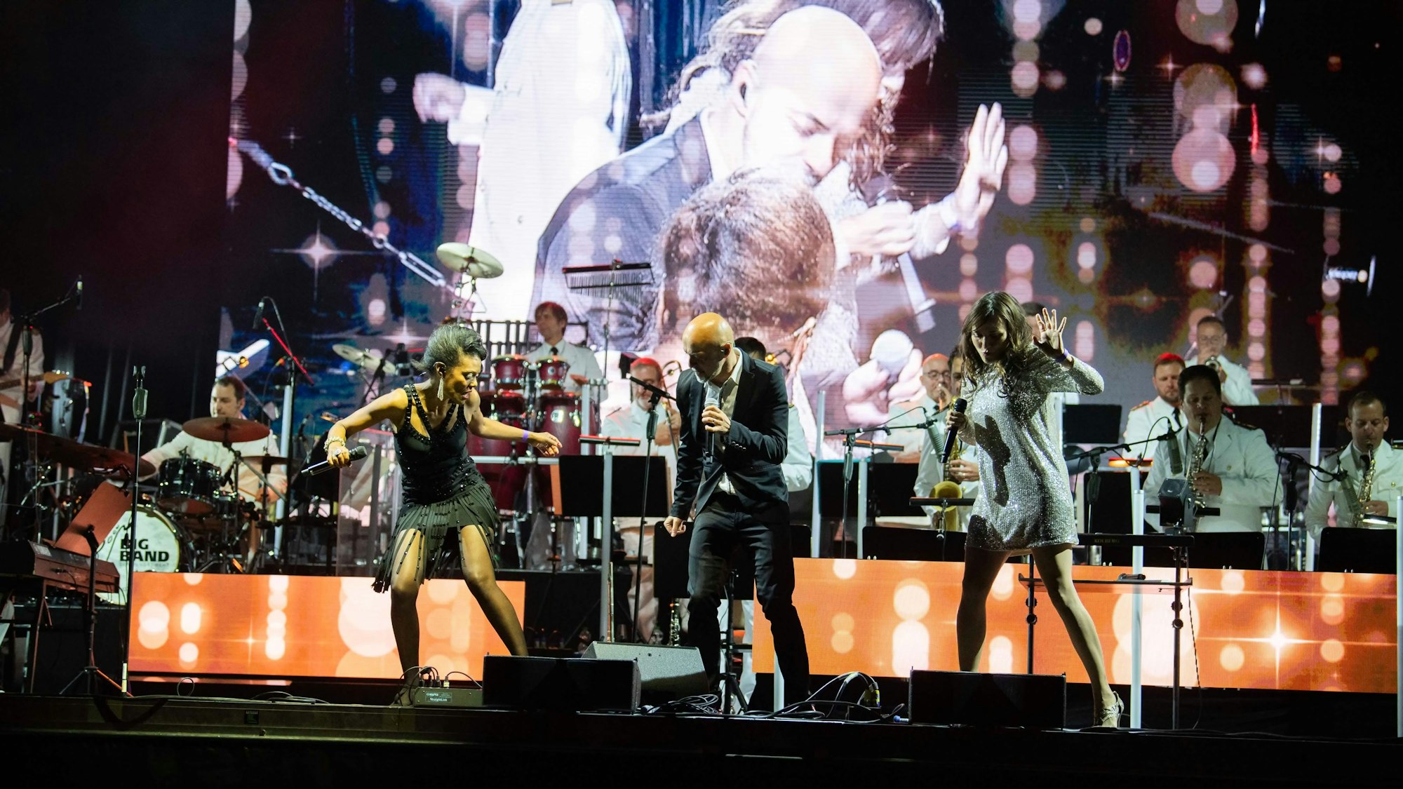 Drei Künstler stehen auf der Bühne mit dem Orchester im Hintergrund.