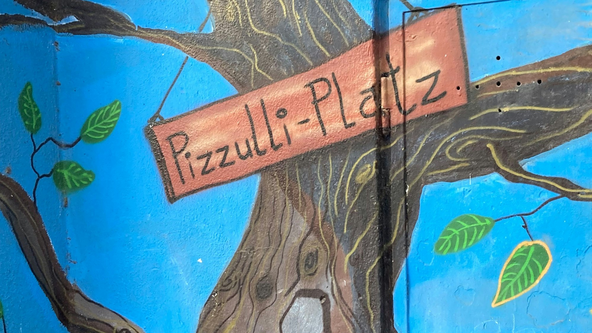 Eine Wand-Deko am Spielplatz Glasstraße, die einen Baum zeigt, an dem ein Schild hängt „Pizzulli-Platz“ ist dem beliebten Streetworker gewidmet