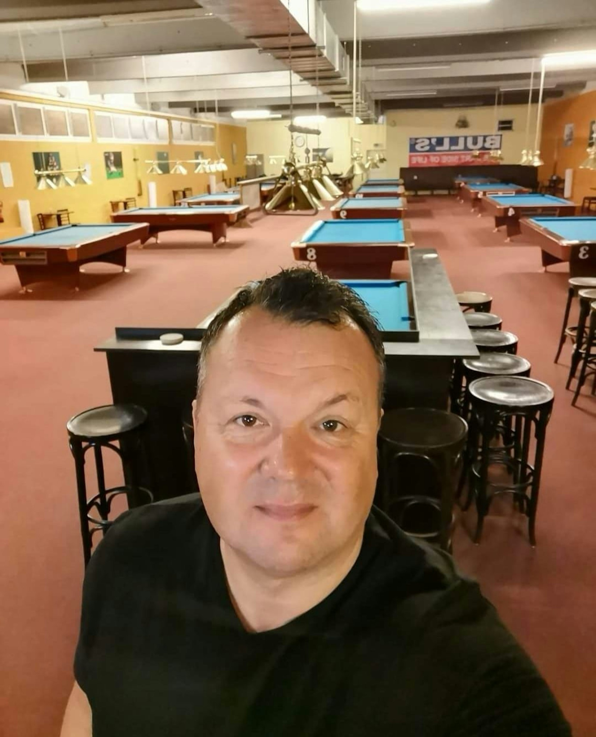 Sascha Müller, der Betreiber des Cologne Cue Club in Köln-Nippes, macht ein Selfie in seinem Laden. Im Hintergrund sieht man viele Billardtische.